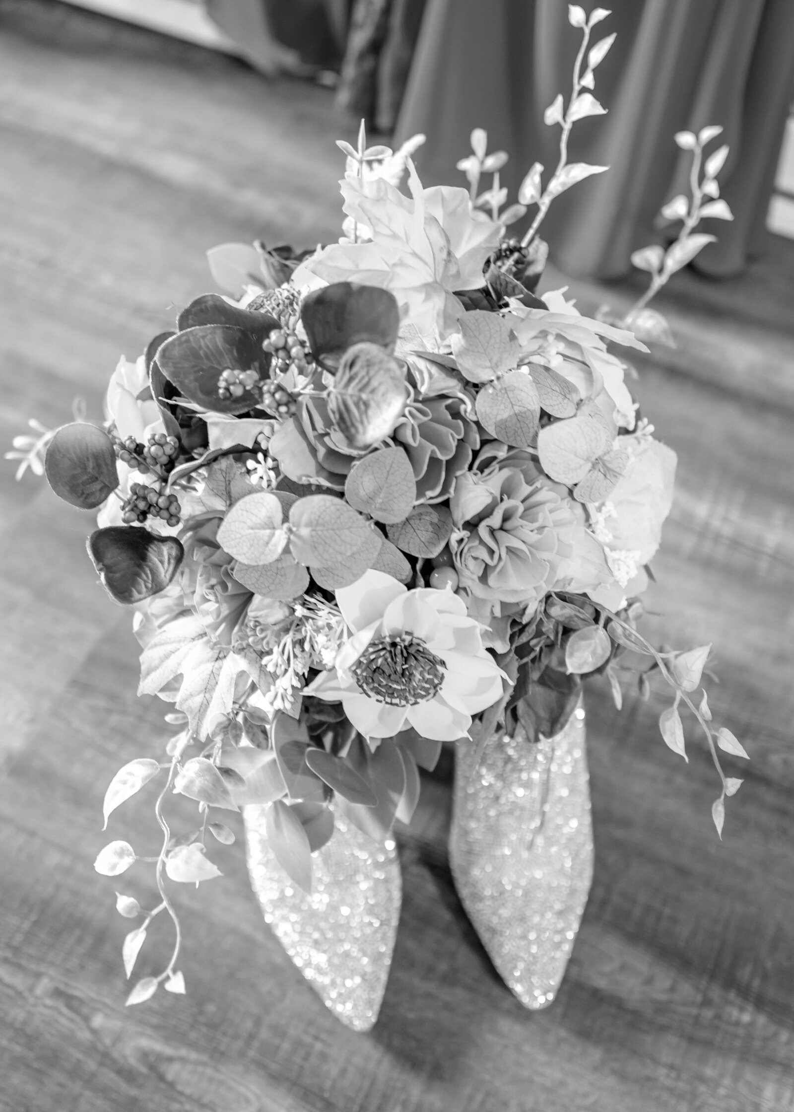 Black-and-white image of a stunning bridal bouquet with lush greenery and textured flowers, placed ion glittering wedding shoes, captured at a Blue Knob Ski Resort, PA wedding, showcasing rustic elegance and high-end wedding details.