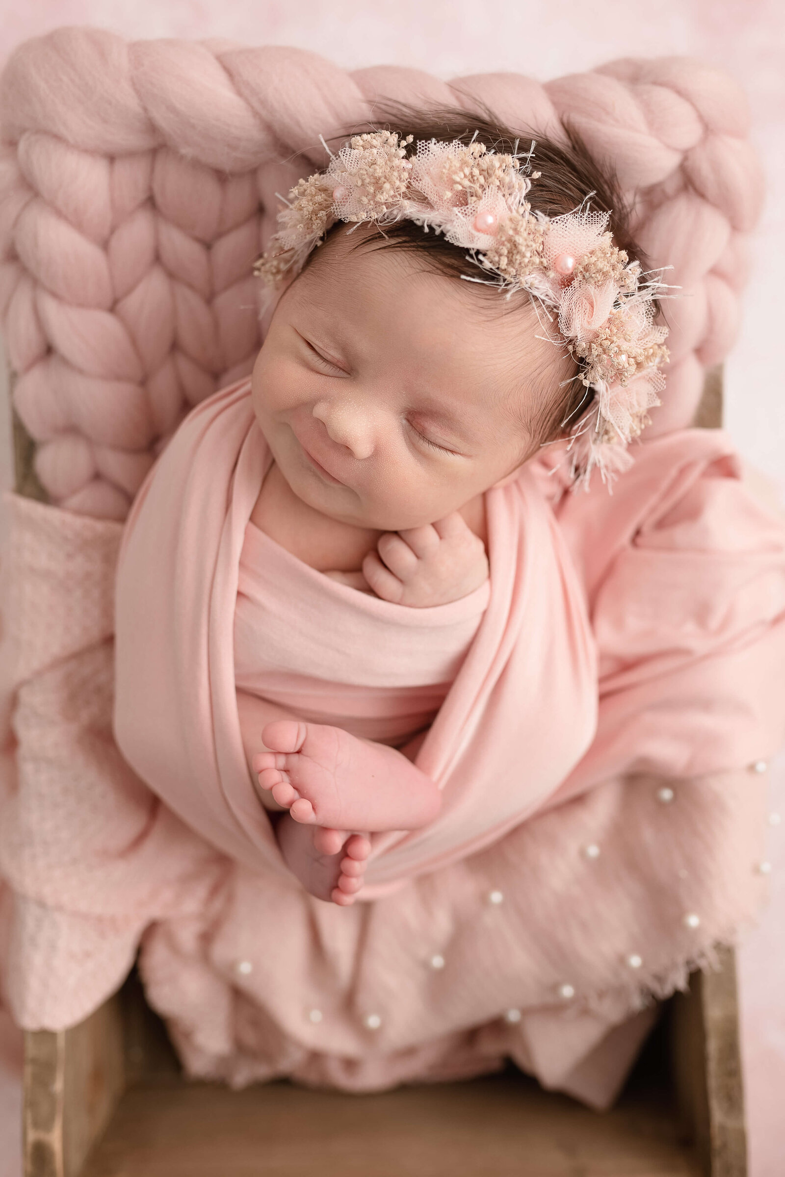 newborn girl wearing a pink swaddle and a pink halo sleeping and smiling in a wooden crate