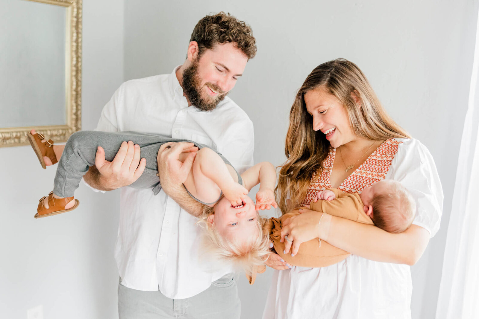 Mom and dad laugh as mom holds newborn and dad holds laughing upside-down toddler