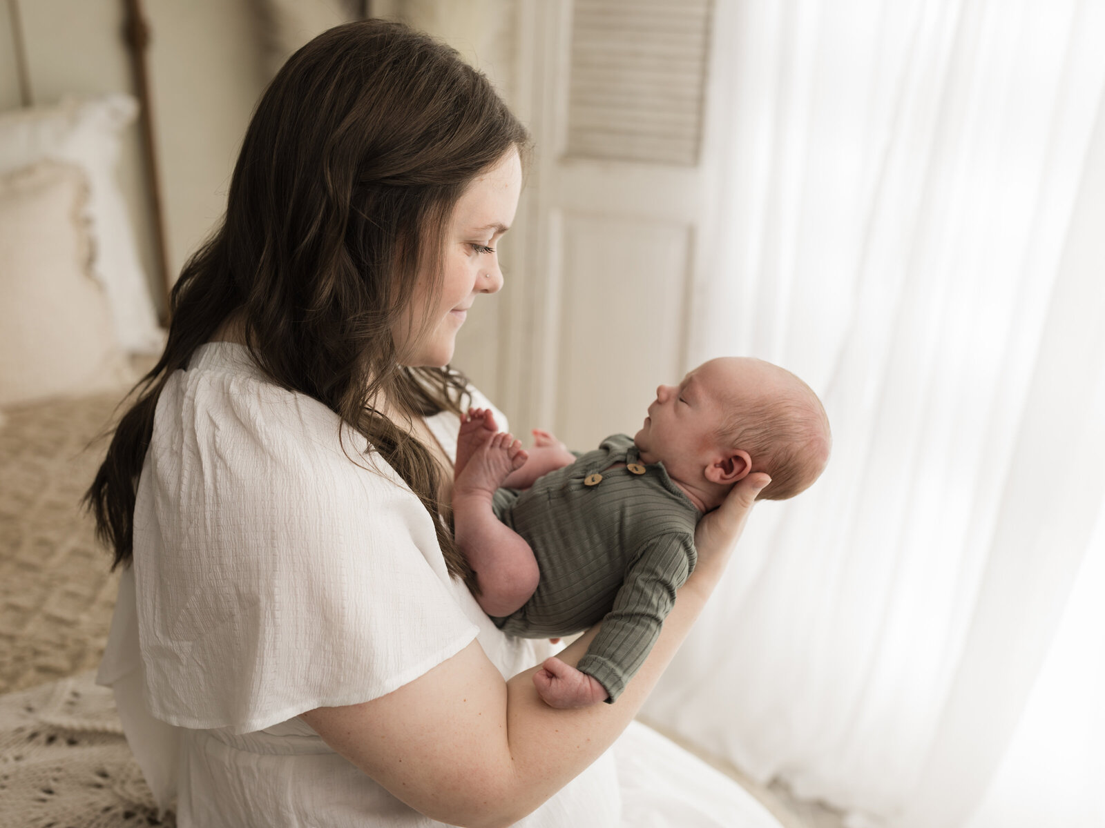 mom in white dress sitting on bed holding newborn baby boy in green onesie cleveland newborn photographer