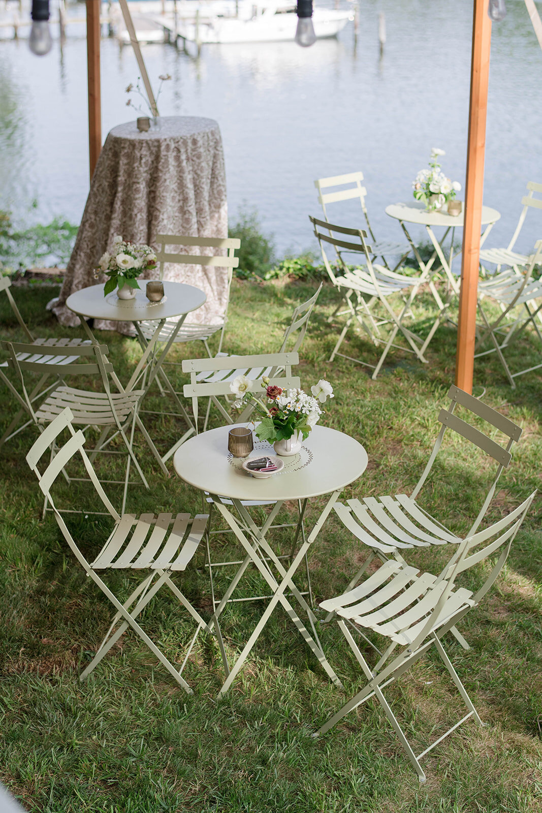 Tented waterfront cocktail hour for micro-wedding with bistro-style seated cocktail tables and high-tops with printed linens. Flower arrangements with anemones and daisies, amber votive candles, and custom matchbooks on cocktail tables.
