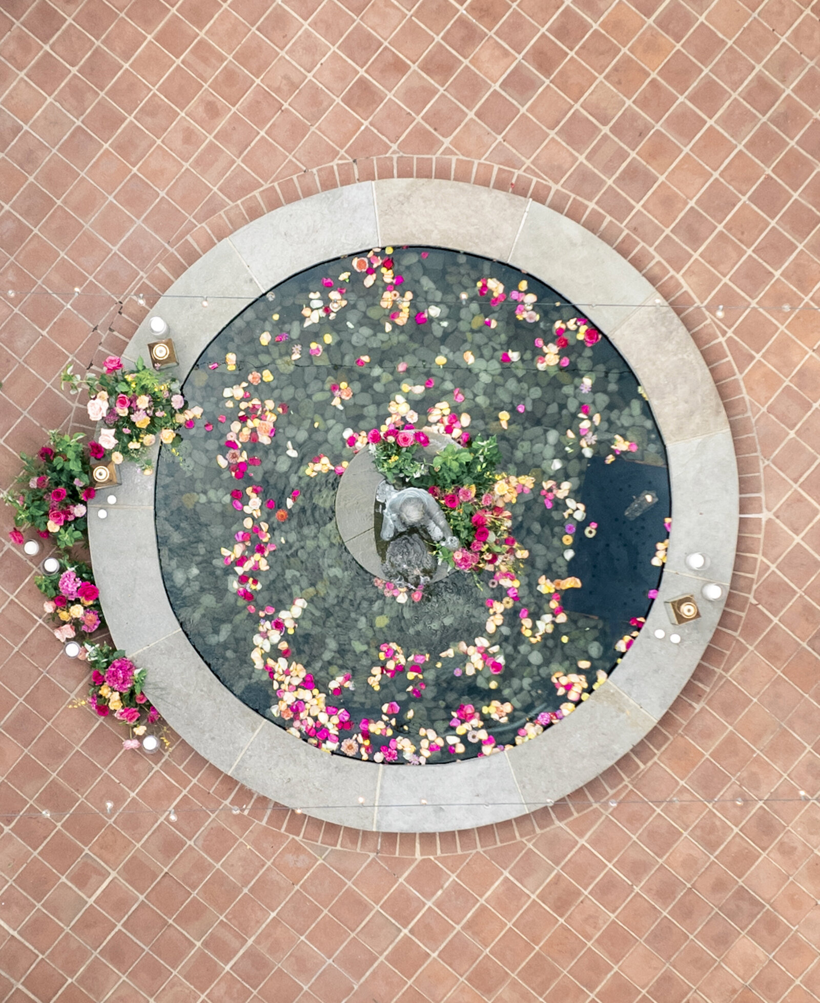 Overhead drone view of fountain at the Gramercy mansion. Bright colored rose petals floating in the water and a floral installation in the middle and side of the fountain with gold lanterns and candles.