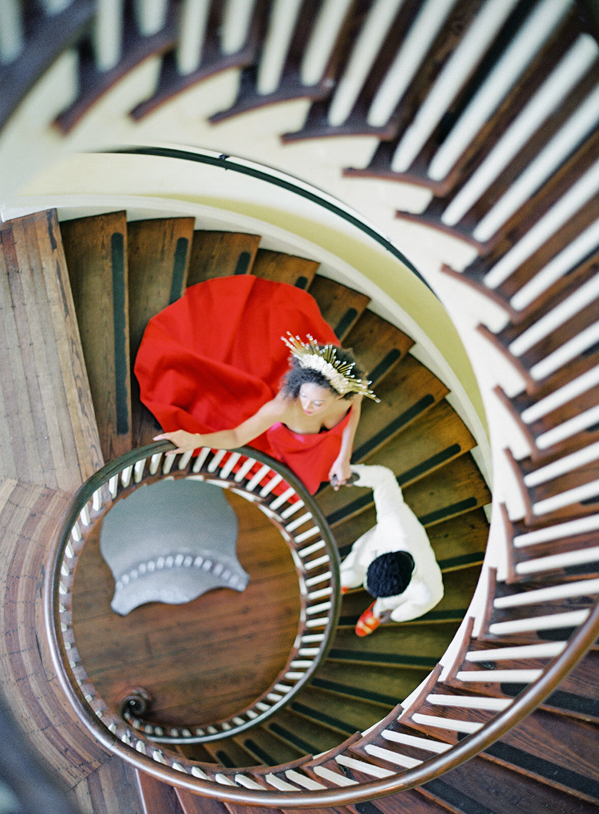 Looking down a spiral wooden staircase with ivory railing. Groom in ivory tuxedo leading bride in red wedding gown and gold sunburst crown down the staircase. Photographed at Lowndes Grove by wedding photographers in Charleston Amy Mulder Photography.