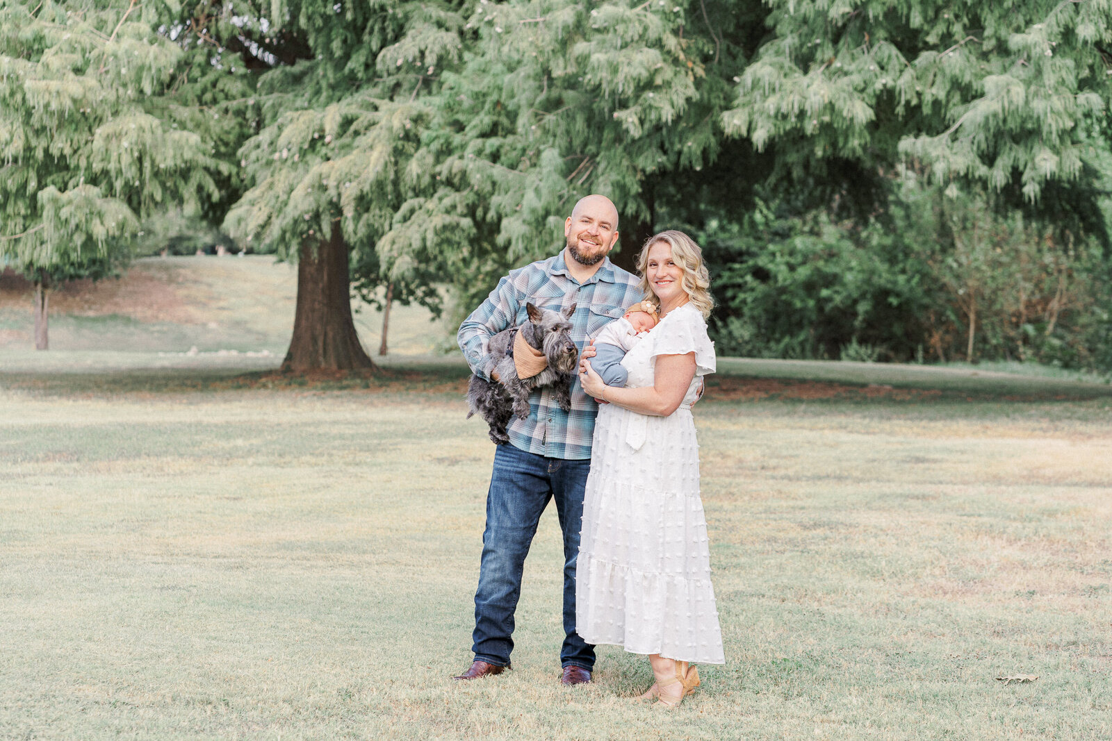 A couple stands in an open field at Katherine Fleischer park. The husband holds their small grey dog while the mother holds their newborn baby.