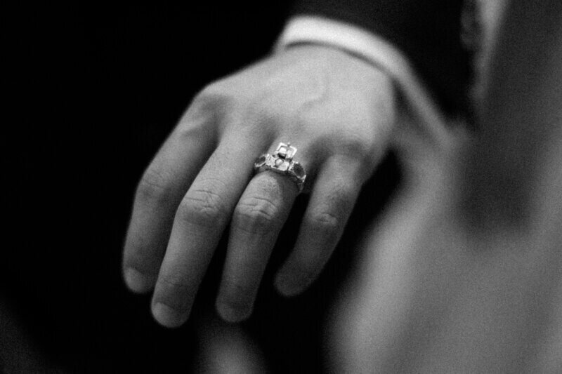 A close-up black and white image of a hand wearing a ring with a large square gemstone on one finger. The background is blurred, emphasizing the elegance of the jewelry.