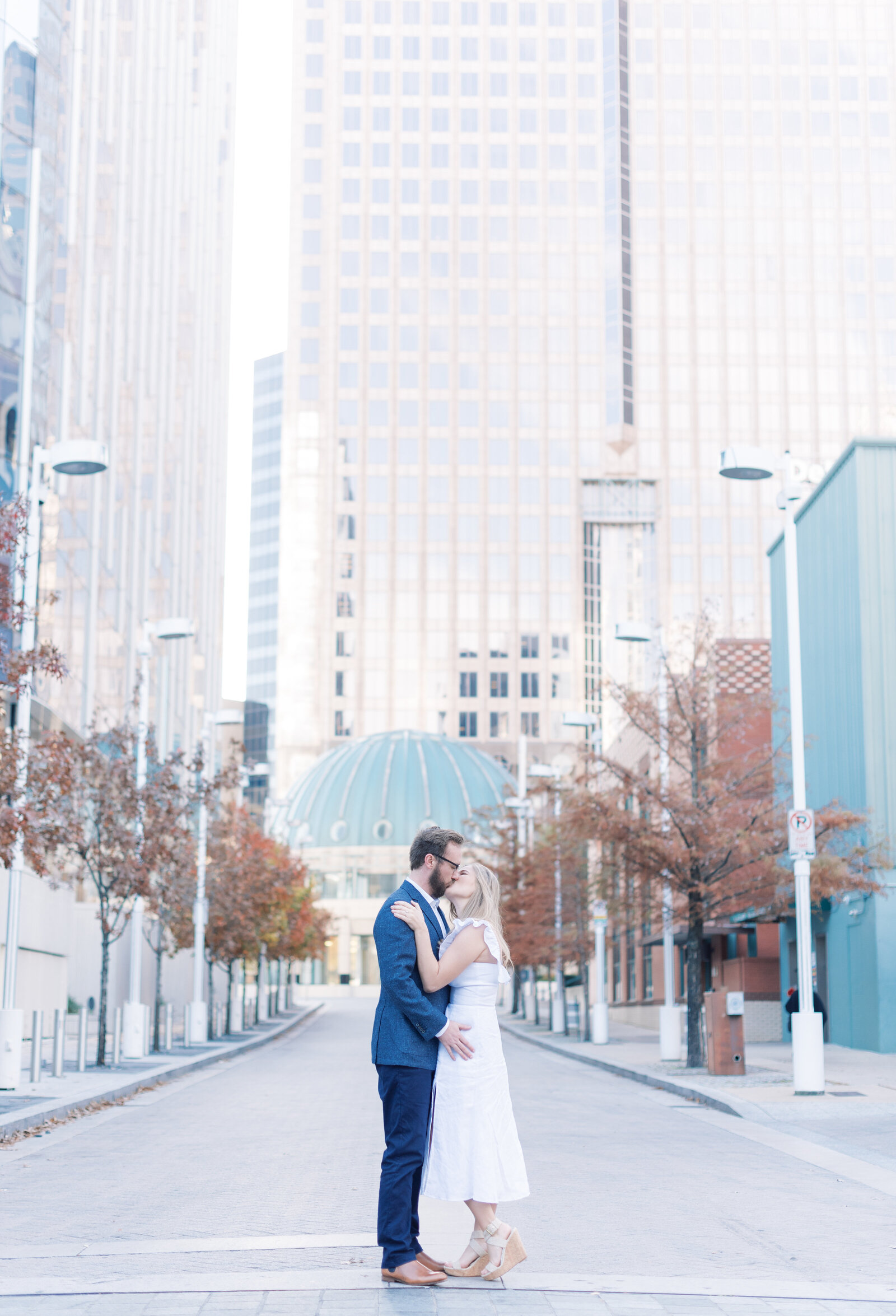 Portrait of a man in a blue suit and a woman in a white dress kissing and holding each other in front of city buildings