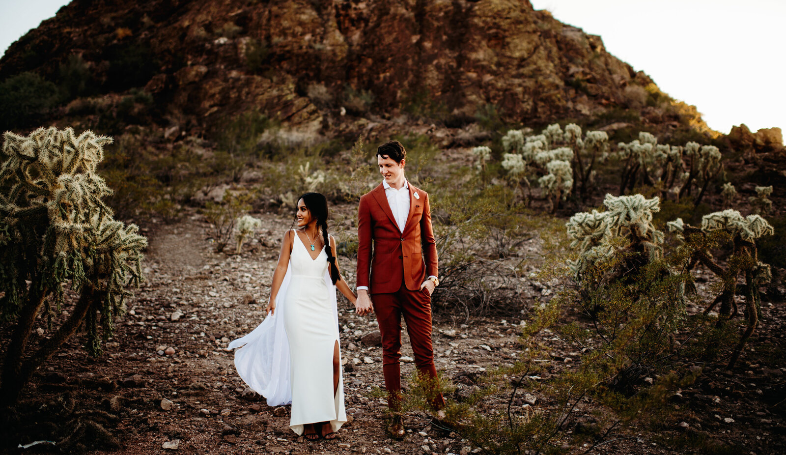 Husband wearing an orange suit holding his wife's hand and smiling at her in the AZ desert. Wife is wearing a fitted white wedding dress with a flowing cape.