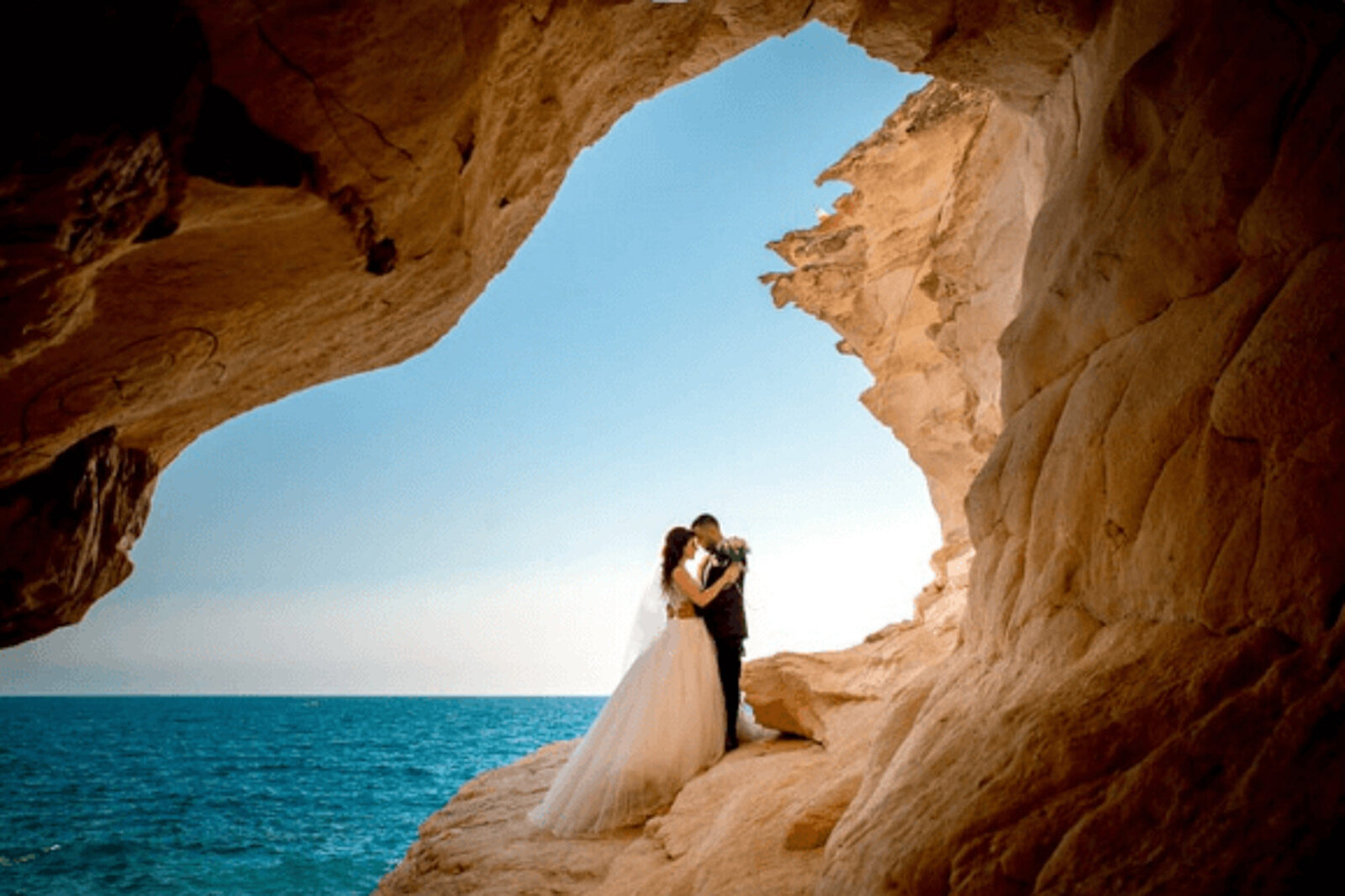 Bride and groom looking at each other under a rock formation on the beach