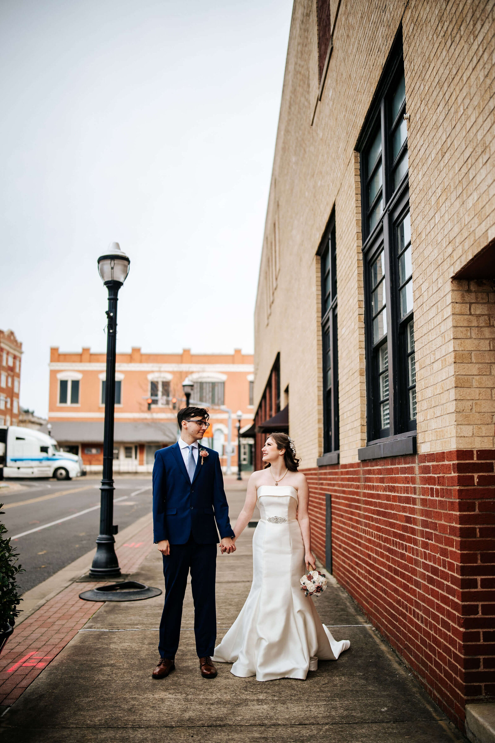 A bride in a white dress and a groom in a blue suit hold hands while walking on a city sidewalk next to a brick building, looking at each other lovingly.