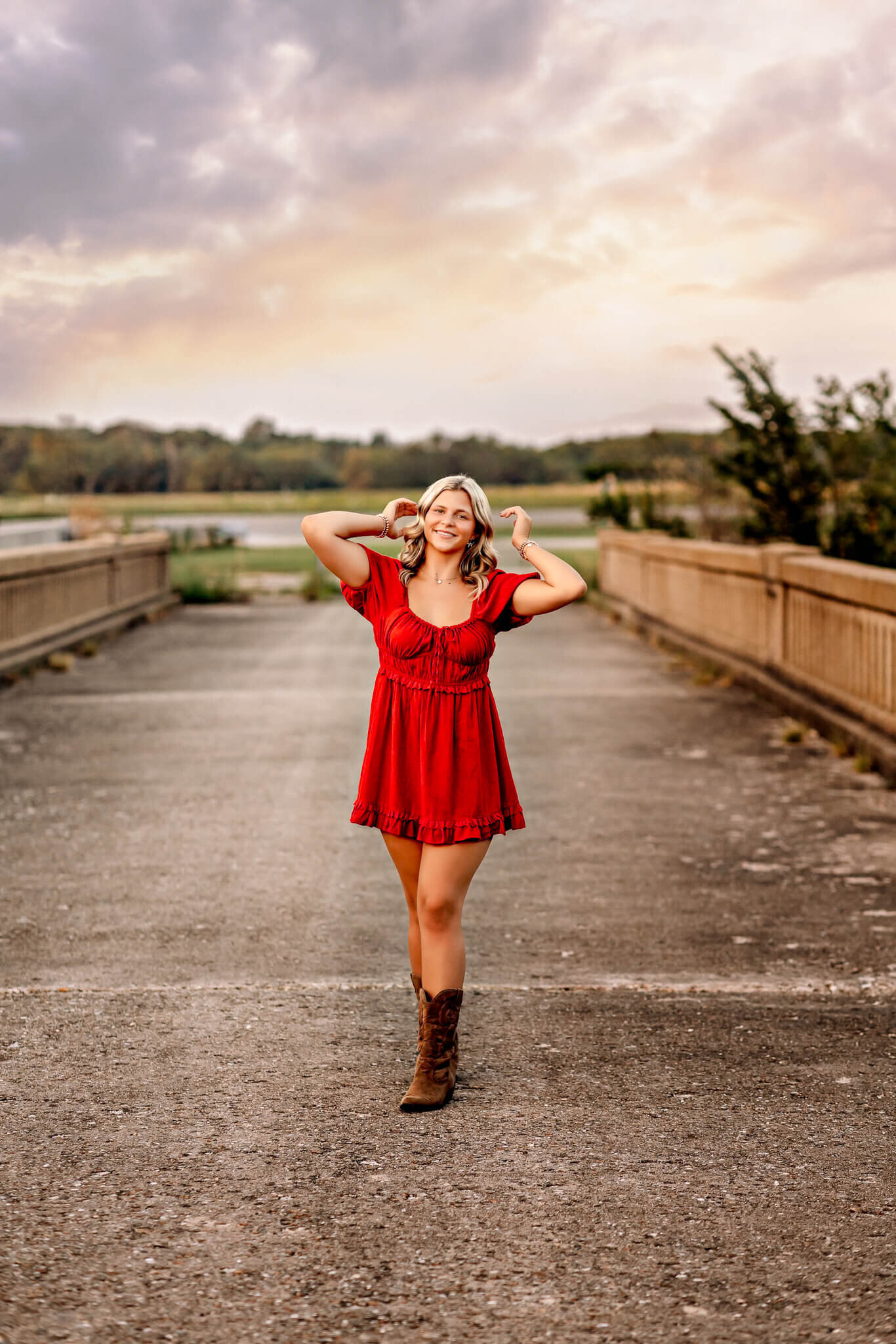 A woman in a red dress and brown boots stands on a bridge, posing with her hands near her face. The sky is partly cloudy, with a soft sunset glow in the background. Trees and greenery frame both sides of the bridge.