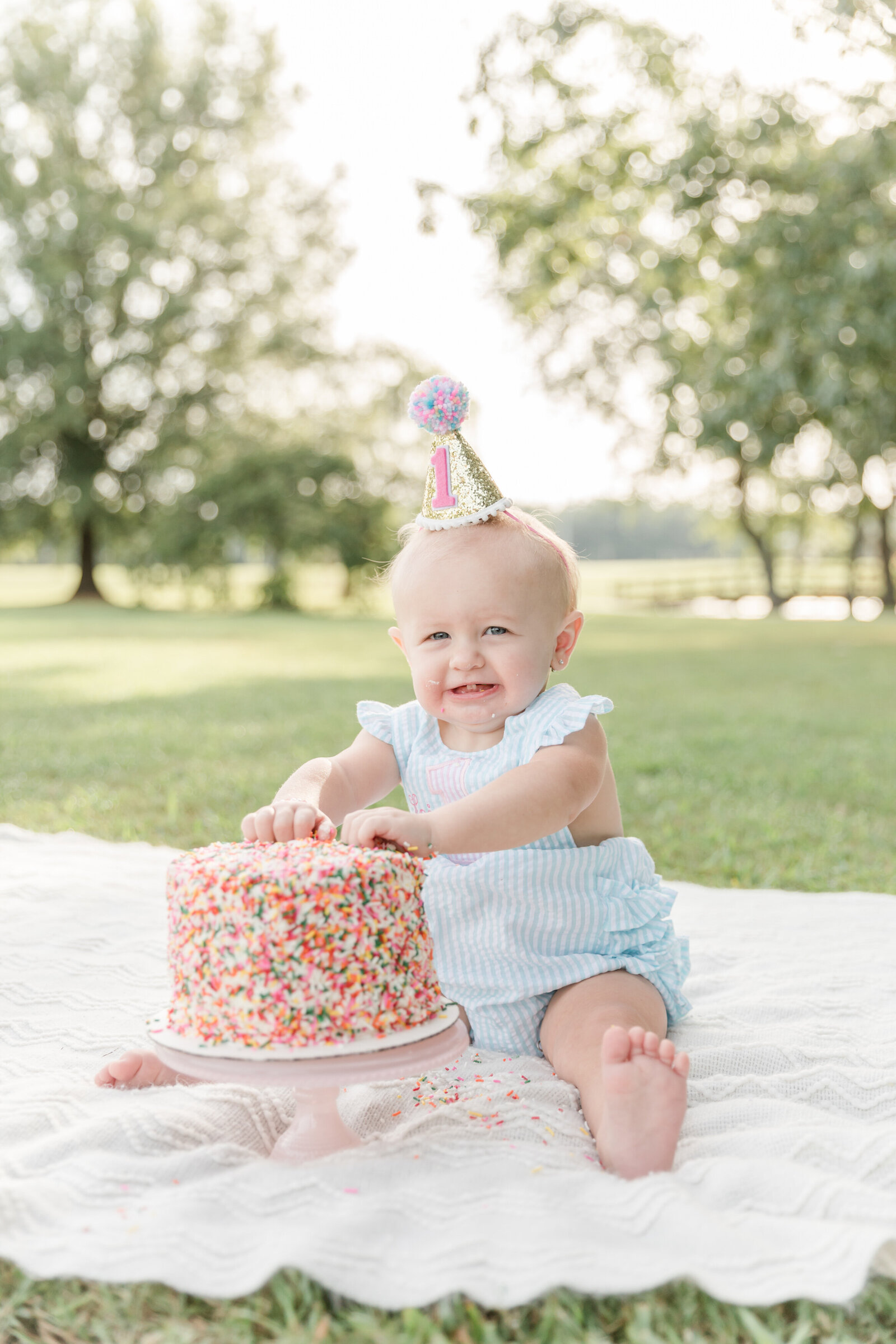 One year old smiling in a party hat while grabbing a sprinkle covered birthday cake.