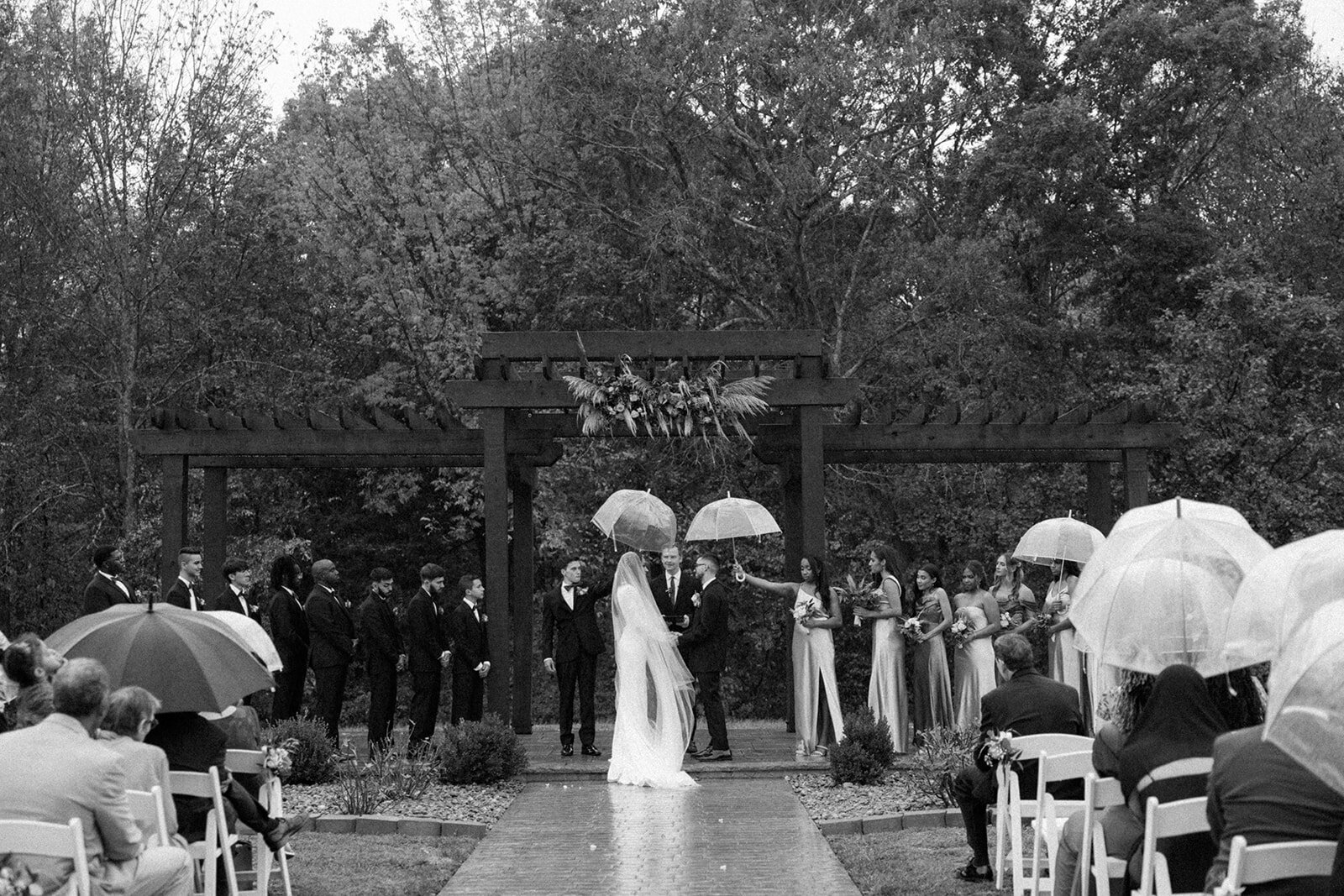 Rain pours over bride and groom on their wedding day and guests hold up umbrellas to avoid getting wet.
