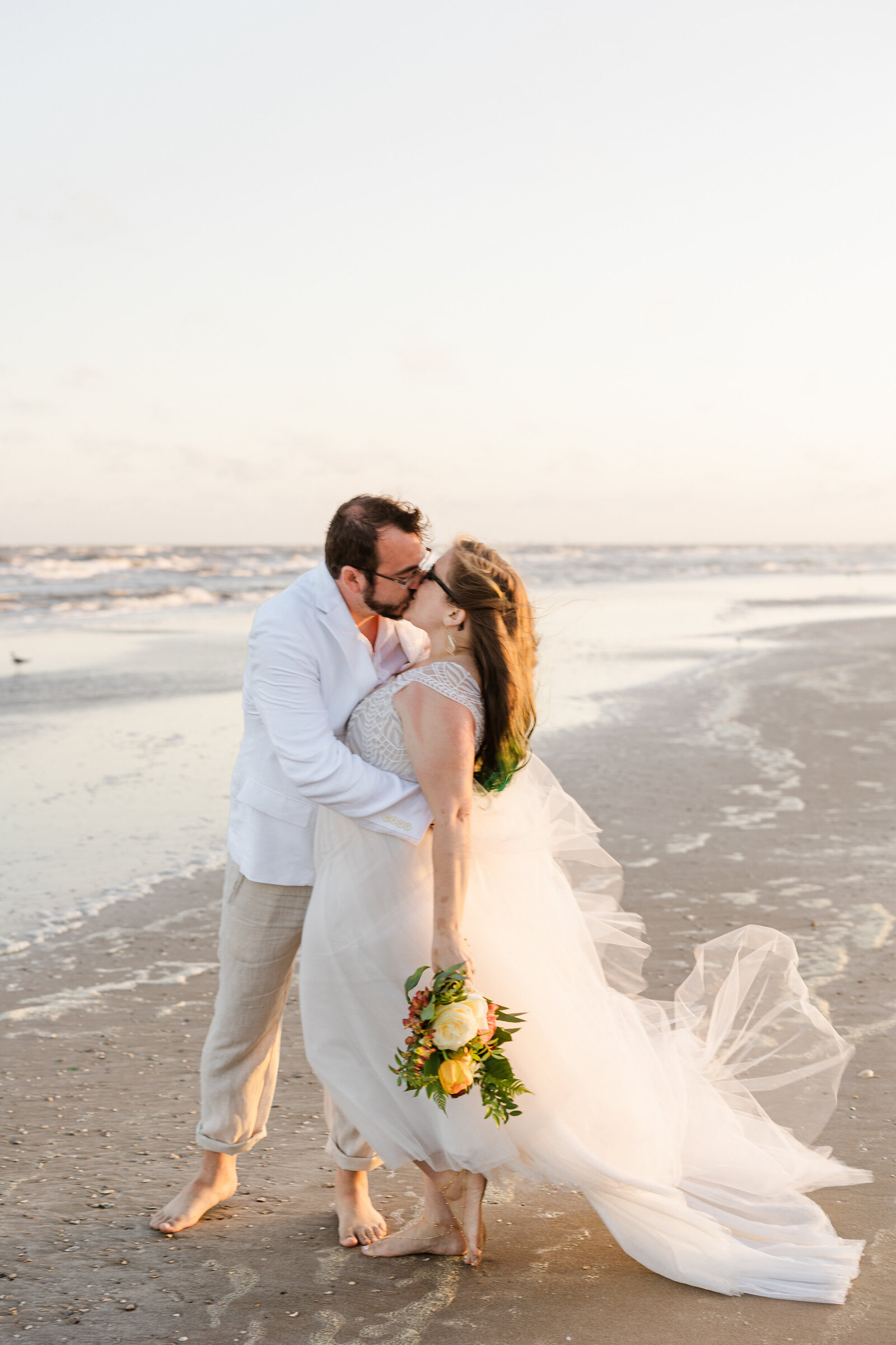 Newlywed couple sharing a kiss on the beach in Crystal Beach, Texas. They are both barefoot and wearing glasses. The bride is on the right and is wearing a flowing white dress and is holding a bouquet at her side. The groom is on the left and is wearing a white shirt and rolled up khaki pants.