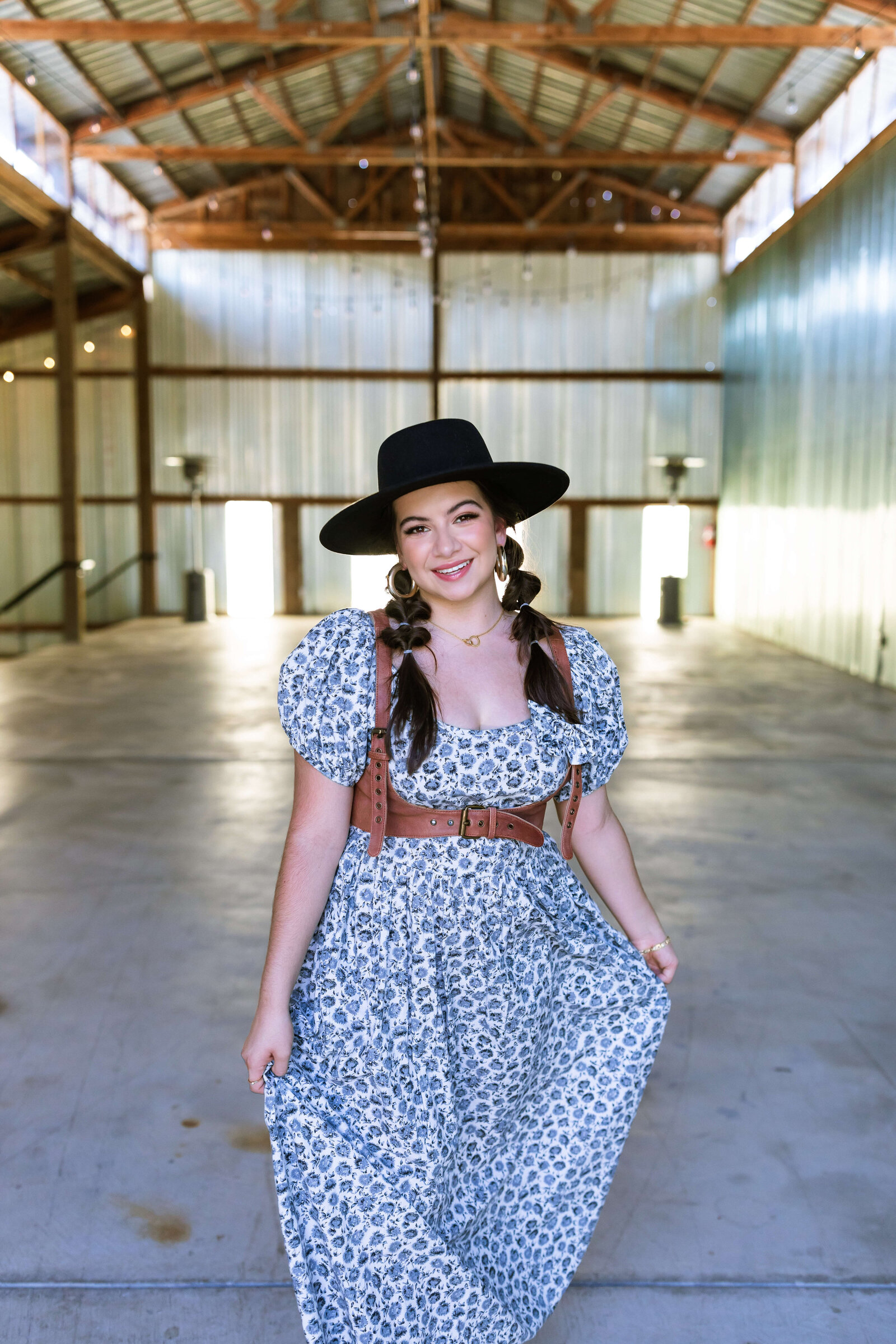 dark haired girl wearing black hat, brown belt, and blue floral dress with bubble braids in a warehouse