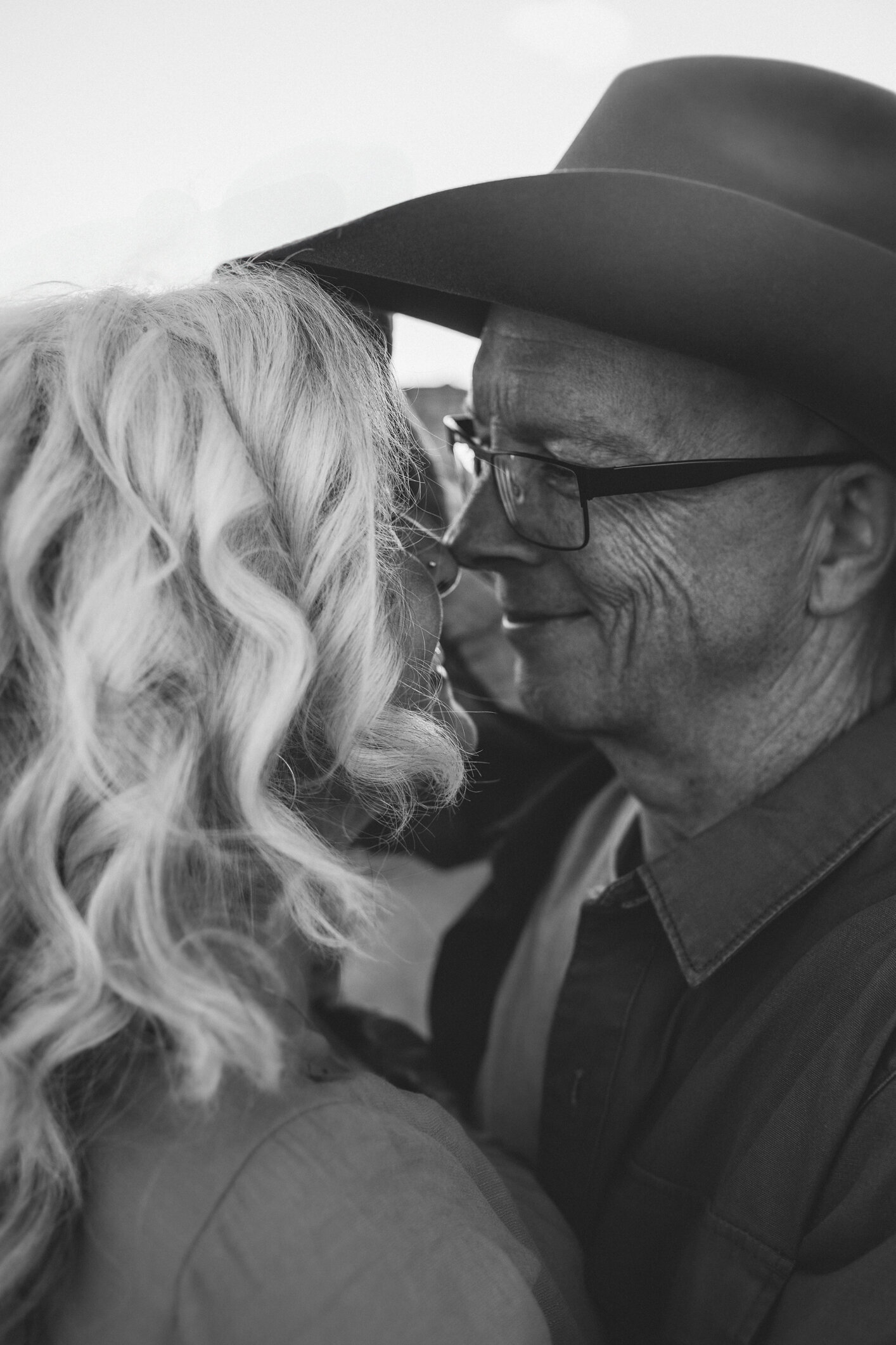 A black-and-white image of a senior husband and wife standing face-to-face, nestling their noses together as the wife lifts up the husbands, cowboy hat so she can reach in for a kiss.