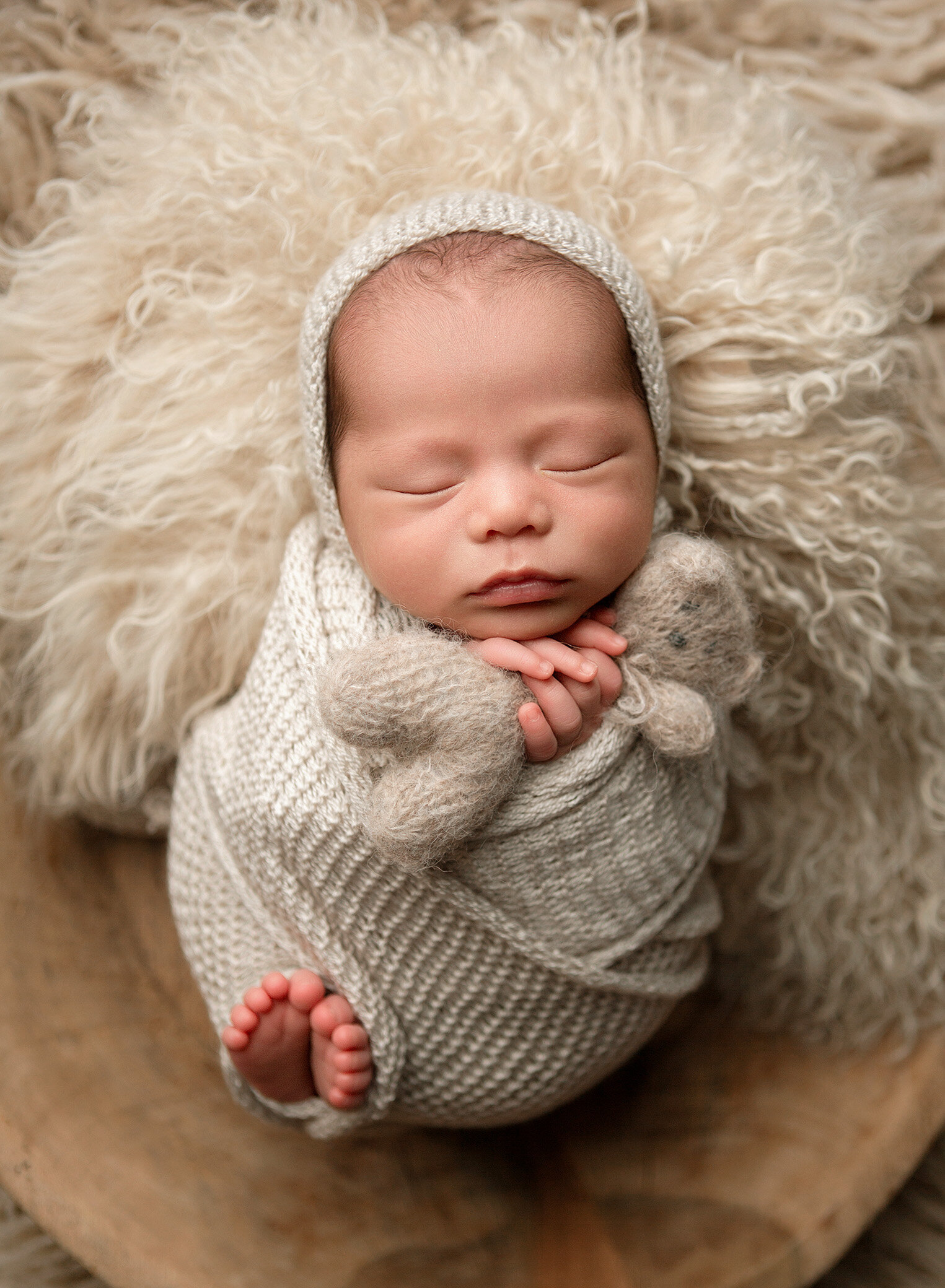 Newborn baby swaddled with his toes peeking out and clutching a tiny knitted bear. He is posed in a vintage wood bowl with soft  fur and wearing a matching knitted bonnet