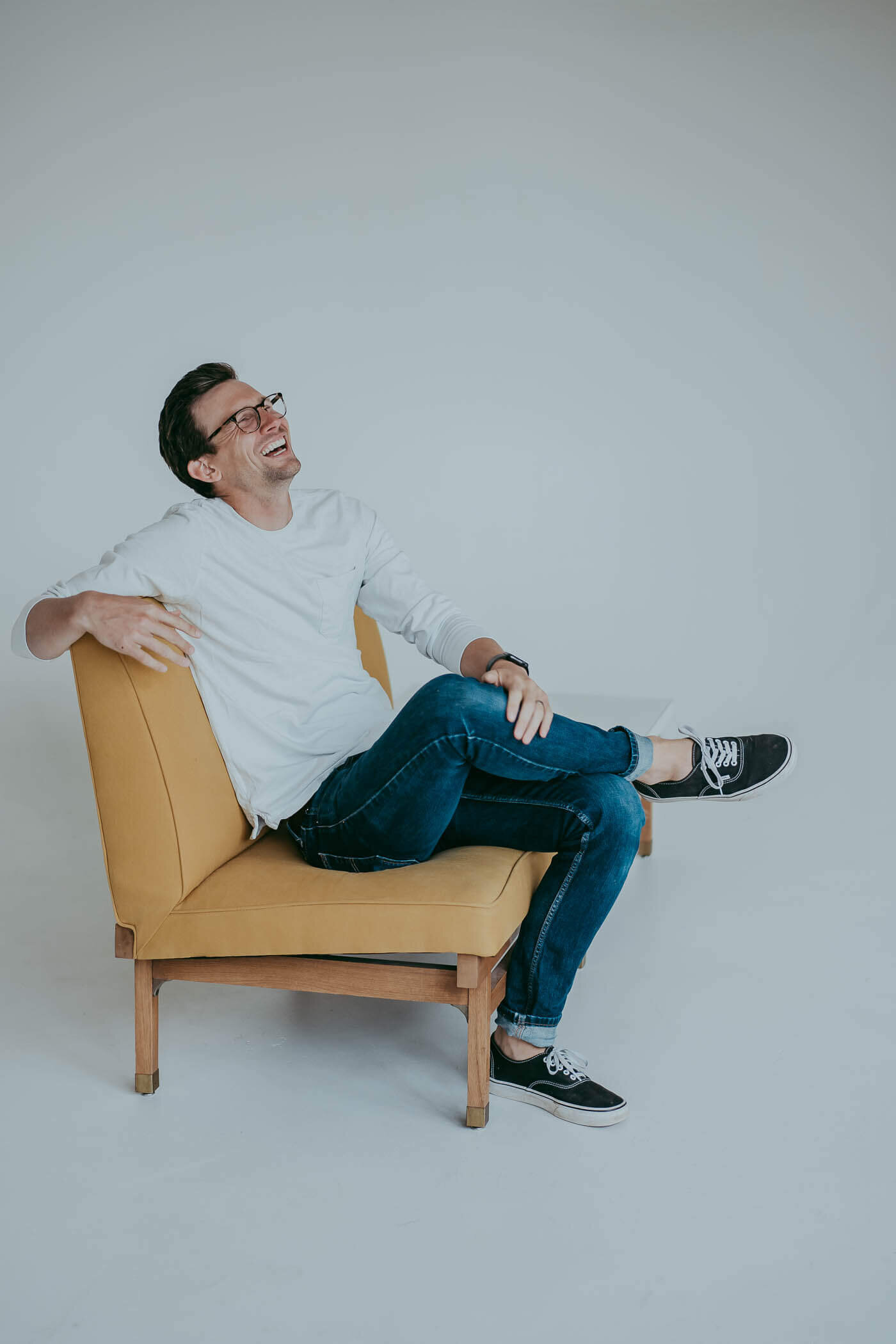 A man sits back in a mustard colored chair and throws his head back, laughing during his personal branding session.