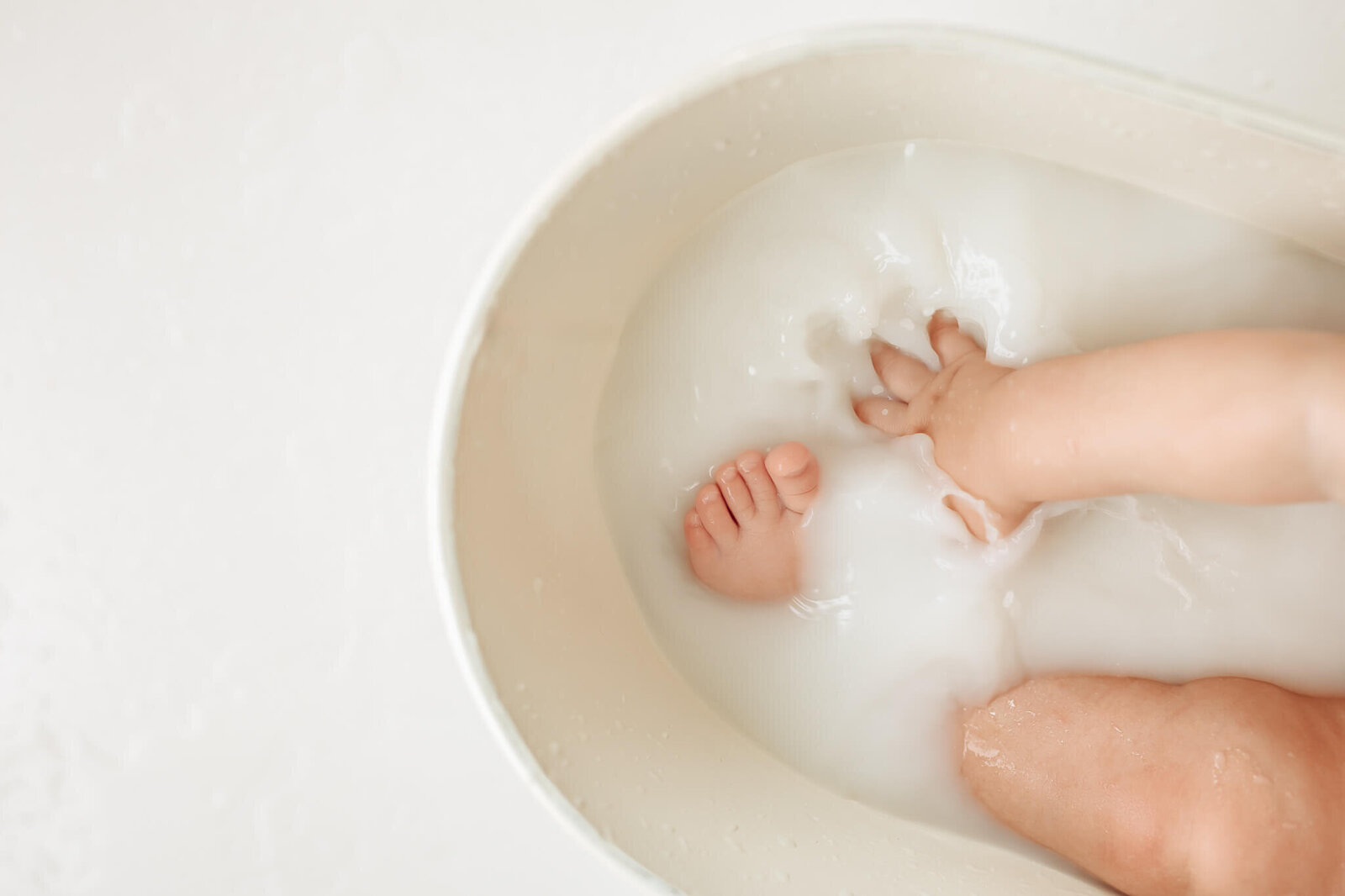 A baby sitting in a small, cream-colored tub filled with milky water. Only one leg and a hand are visible, playfully splashing. The surrounding area is light and minimalistic.