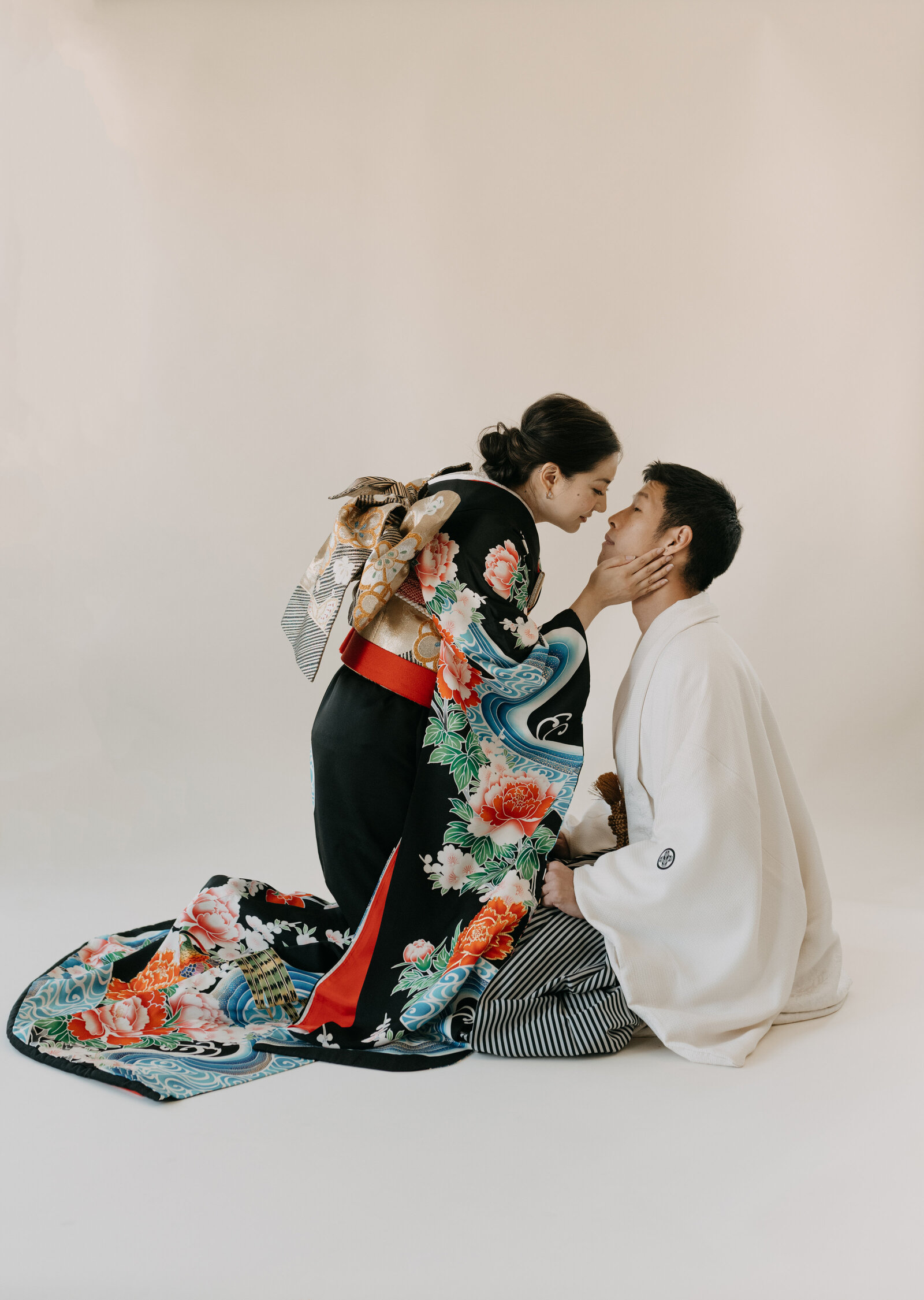 Bride in black peony wedding kimono rental gently kissing groom’s face as he kneels in a Los Angeles California studio | 黒引き振袖レンタル 豪華牡丹柄 花嫁が新郎にキスするスタジオウェディング