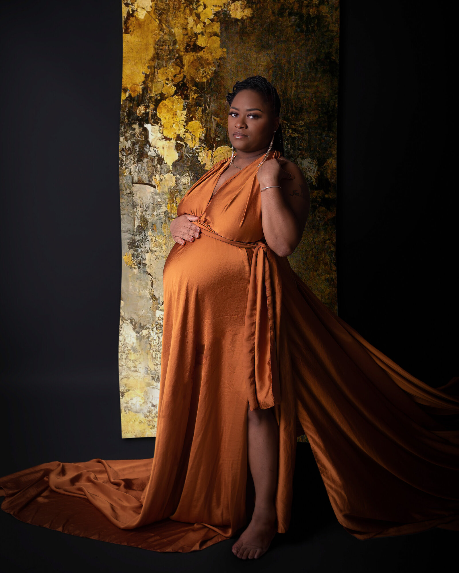 A mother-to-be in an elegant copper dress stands confidently in front of a gold abstract backdrop, her gaze serene and poised.