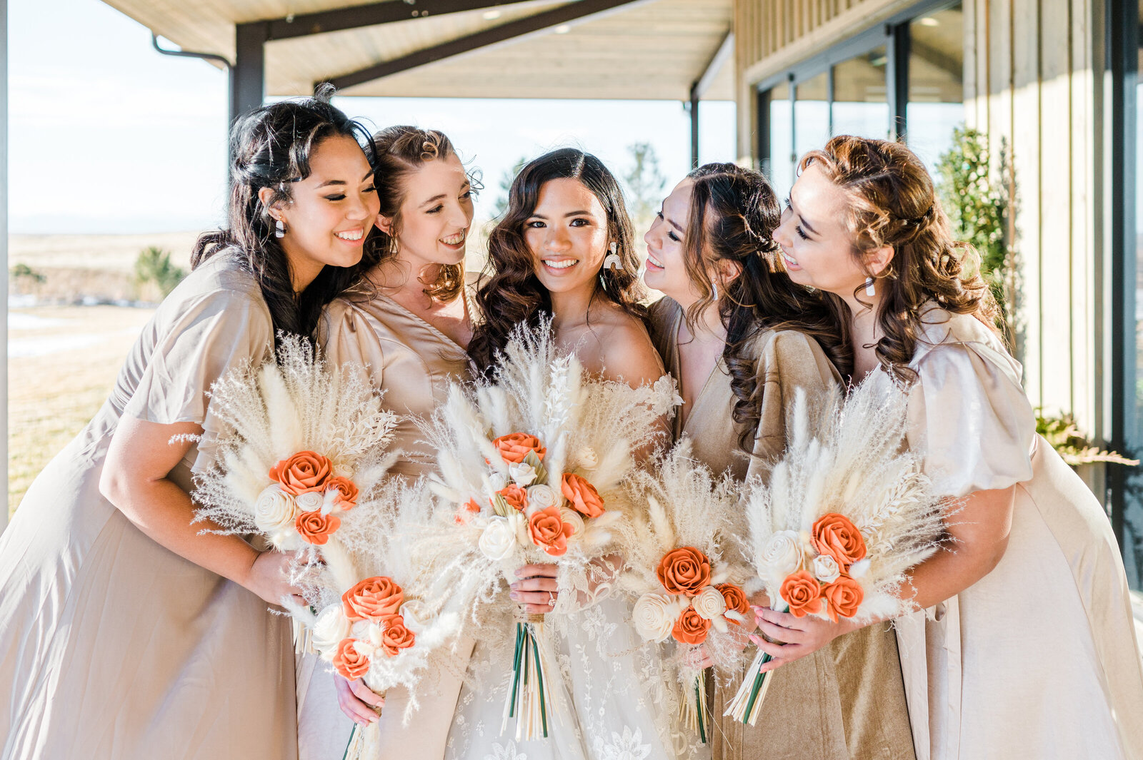 Bridal Party in beige chiffon dresses smiling together and looking at their bride while holding orange and beige flowers