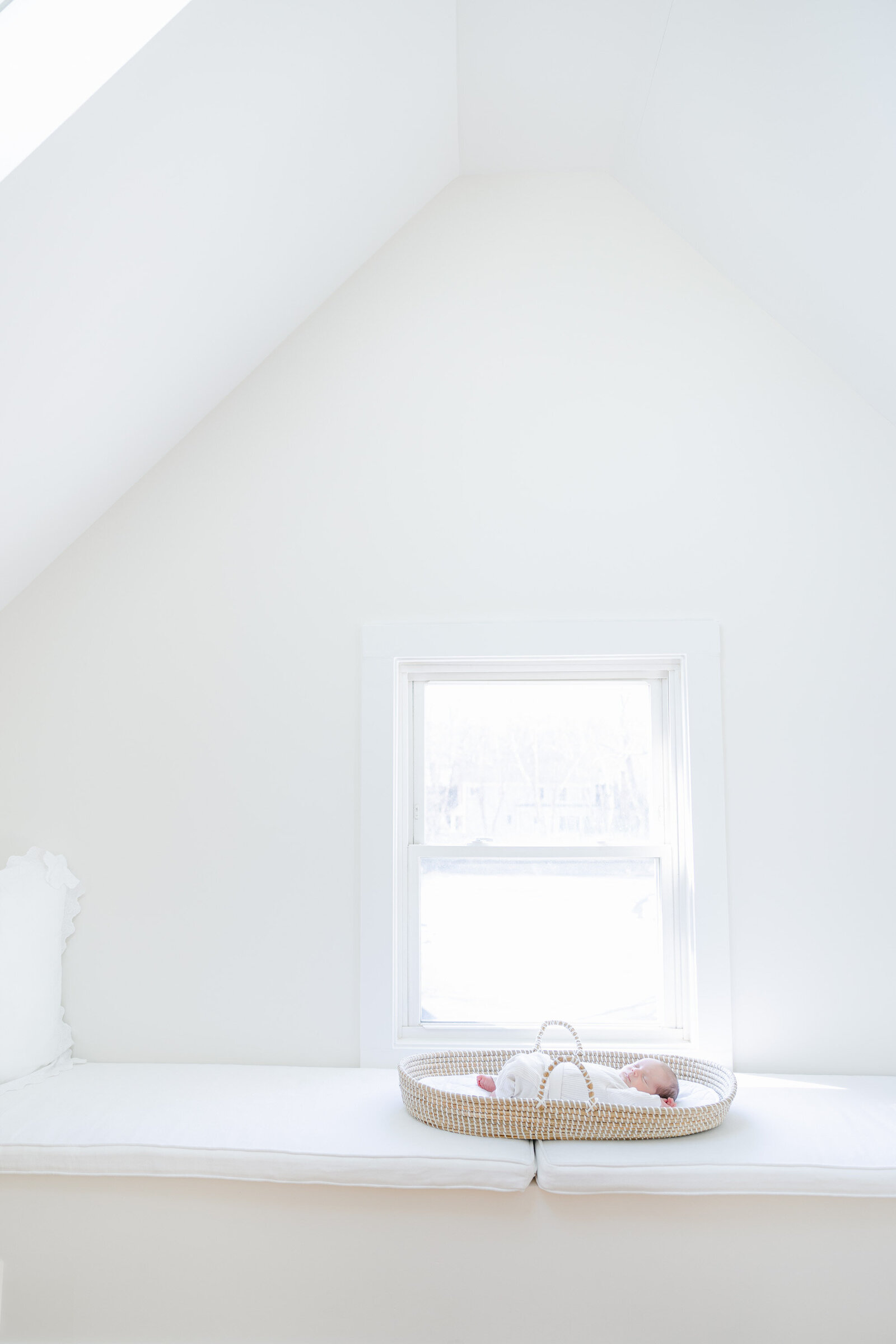 Newborn baby sleeps peacefully in a Moses basket on a white window bench during a bright and airy lifestyle newborn photography session in Fairfield County, Connecticut. The minimalist scene highlights natural light and a calm, serene atmosphere, photographed by Nicole Neira.