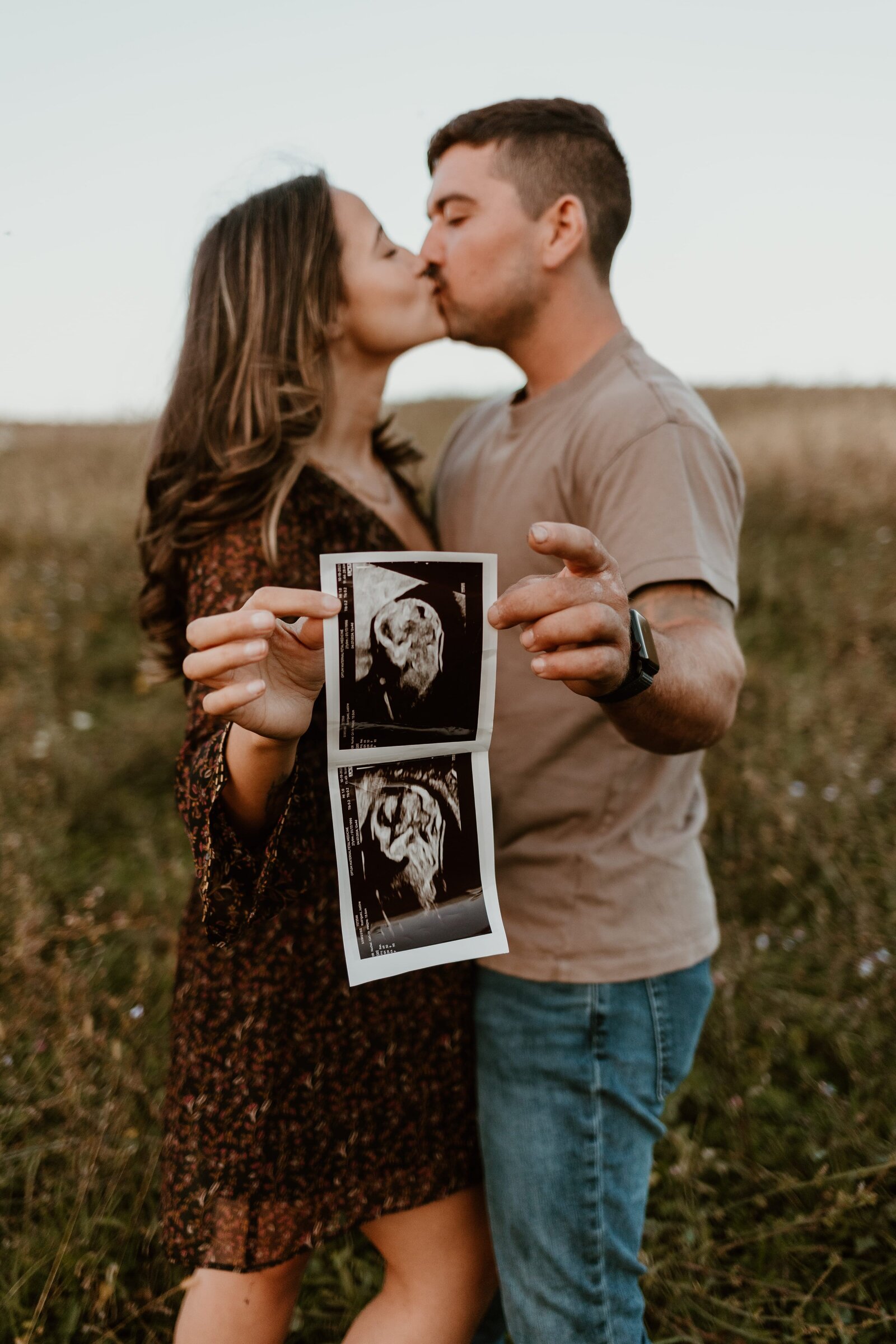 A loving couple stands in a field, sharing a kiss. The woman, dressed in a patterned dress, and the man, in a casual t-shirt and jeans, hold ultrasound images of their baby. The affectionate pose and natural backdrop convey a sense of anticipation and joy for their future together as parents.