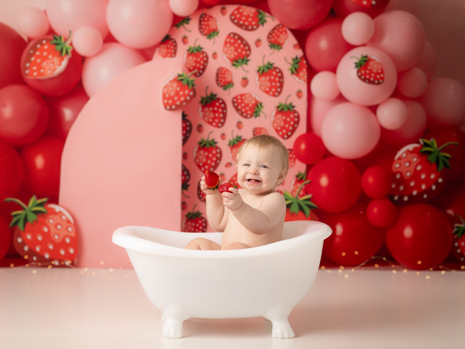one year old girl sitting in tub eating strawberries for first birthday photoshoot