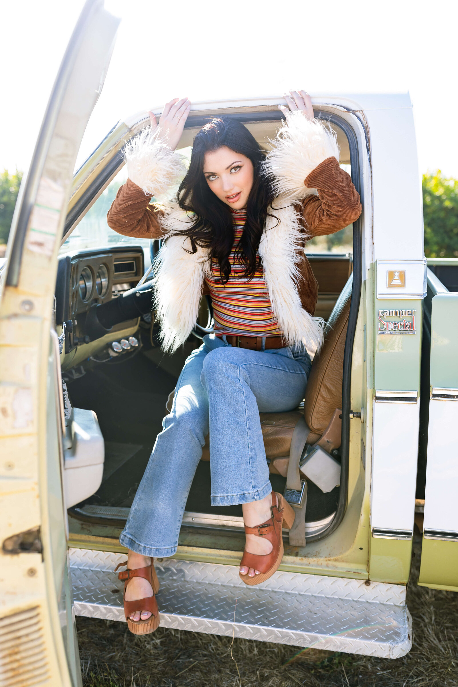 girl with dark hair wearing 90's inpspired outfit with fur coat, striped shirt, and brown wedged heels sitting in an old vintage green and white truck