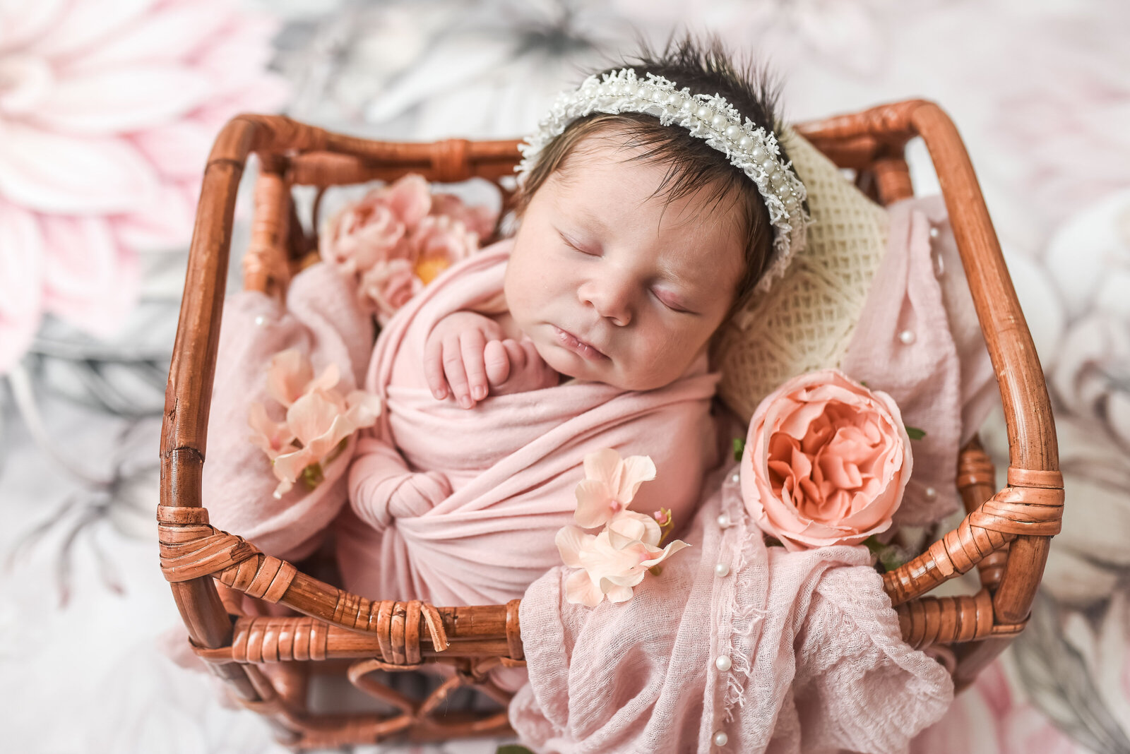 newborn baby sitting in wicker basket wrapped in pink blanket with flowers for photo shoot