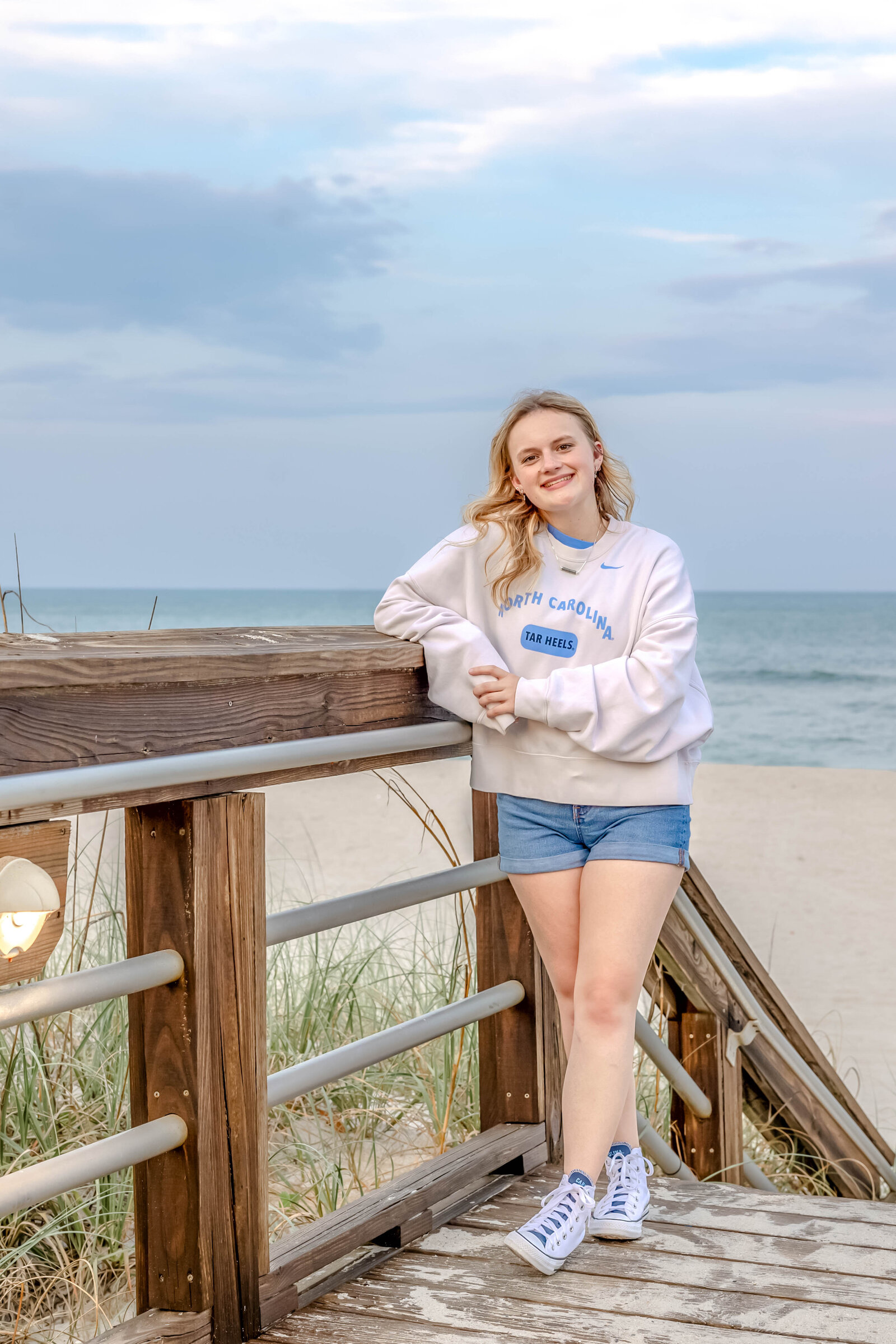 A high school senior in a University of North Carolina sweatshirt and shorts leaning on a boardwalk railing with the ocean in the background in Carolina Beach, North Carolina. This relaxed portrait captures the casual and natural vibe of senior photography, ideal for beach-themed sessions.