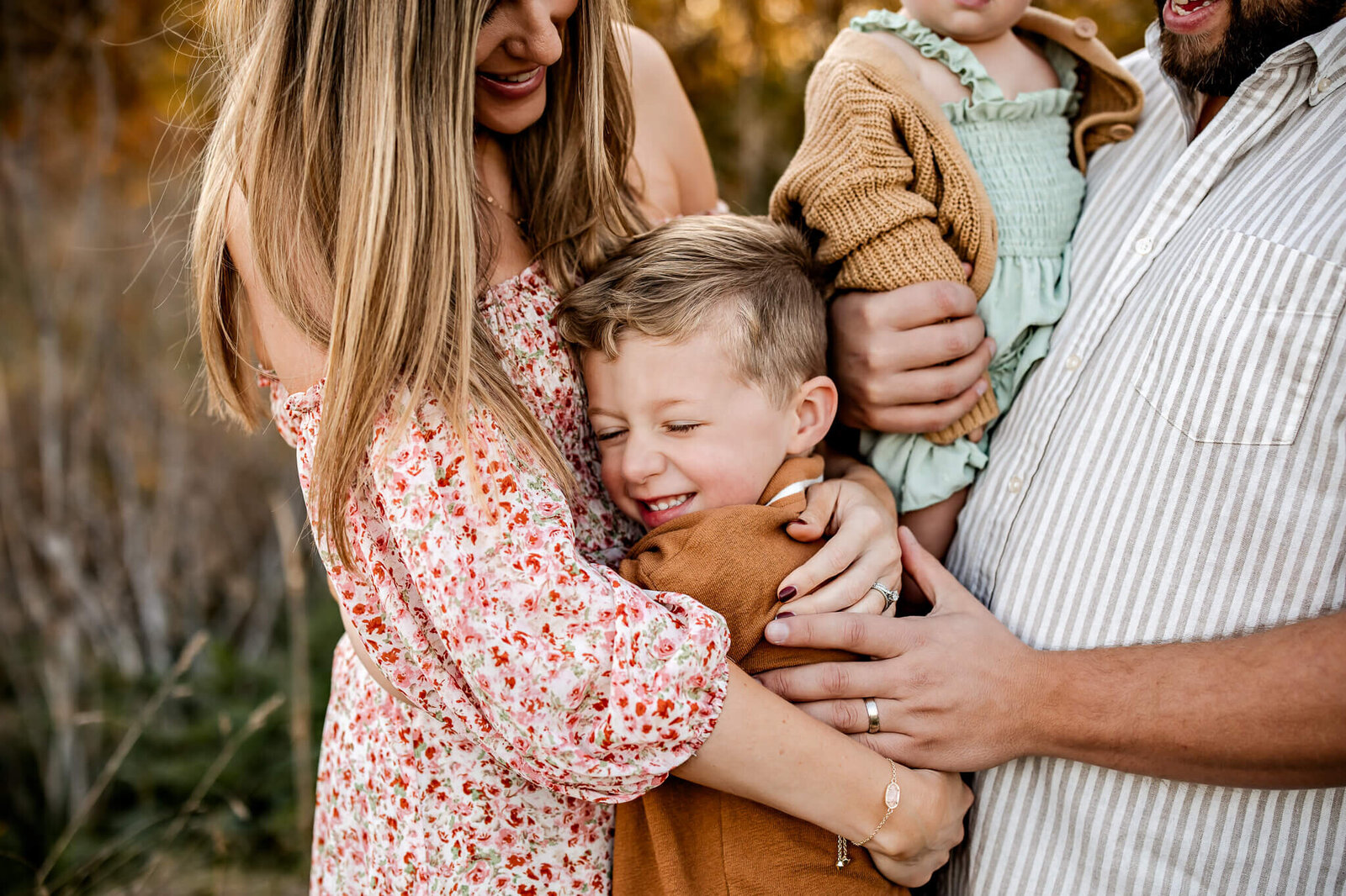 A Bloomington family photographer captures a tender moment as a woman hugs a smiling young boy in a brown jacket. Nearby, another person cradles a baby in green and brown. The outdoor portraits exude warmth against the lush, autumnal backdrop.