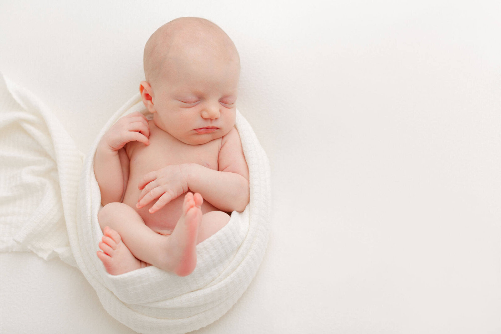 Baby sleeping on back on a soft cream colored backdrop. Baby has a white textured blanket wrapped around her body with her belly, arms, and legs showing.
