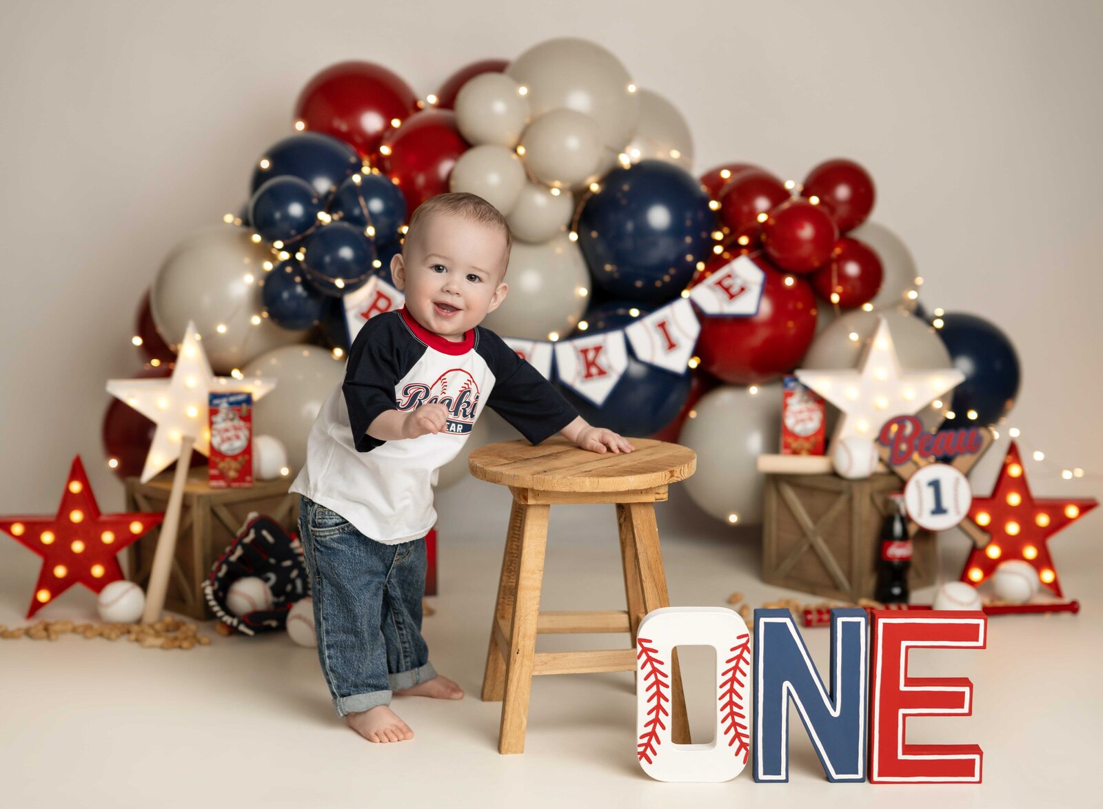 Baby boy standing by stool in front of baseball balloon garland. Cake Smash photo taken by Jenni Nikki in Atlanta, GA