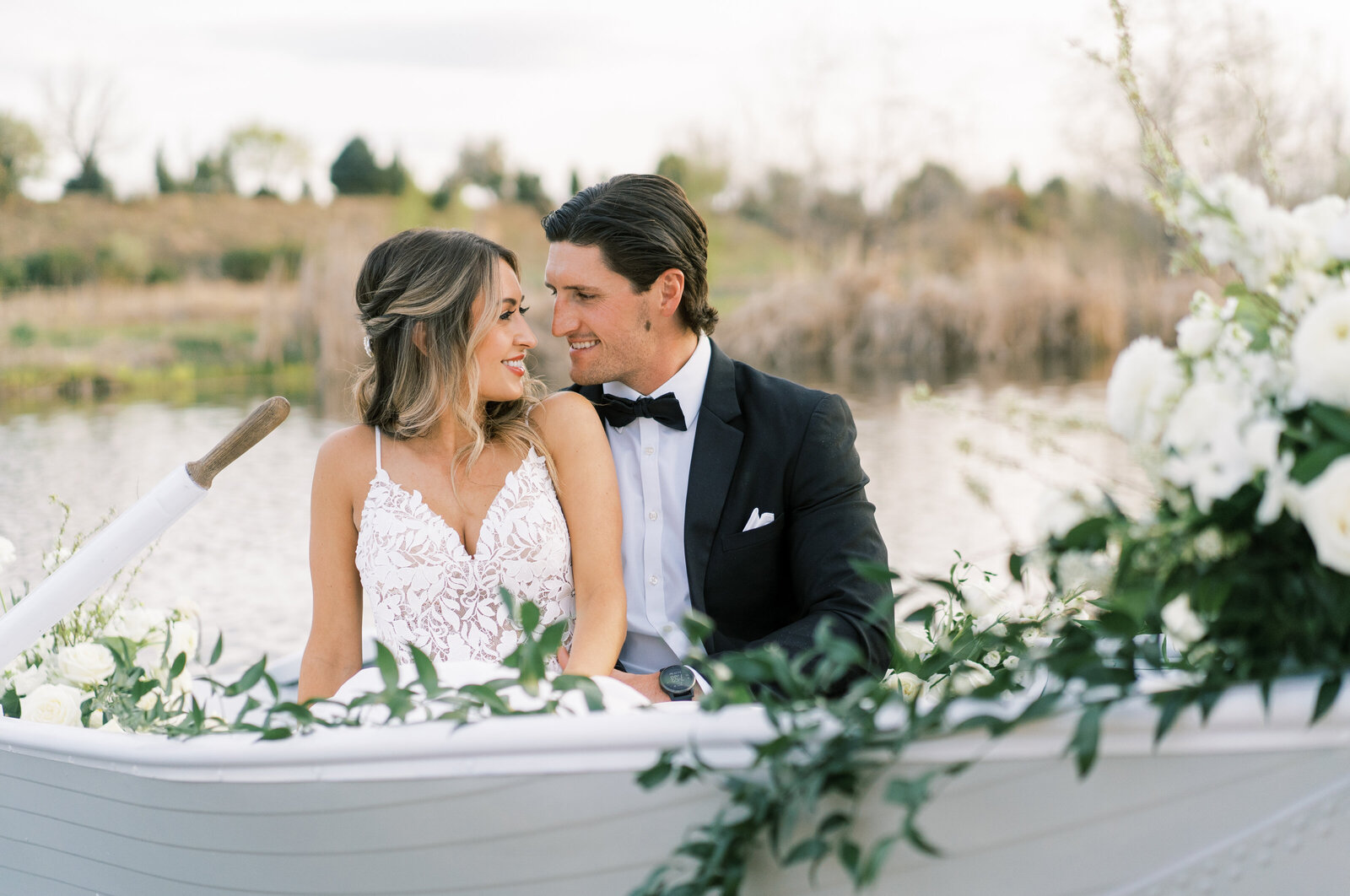 Portrait of a bride and groom in a white wedding gown and black tuxedo in a boat filled with flowers and greenery on a lake