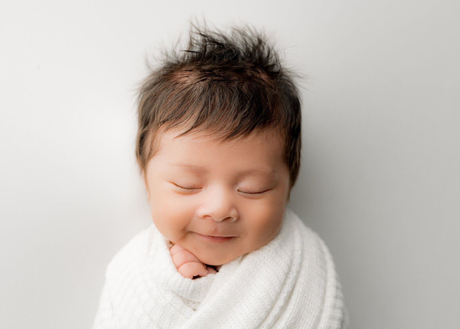 baby boy with lots of hair sleeping on a white blanket, wrapped in a white swaddle and smiling