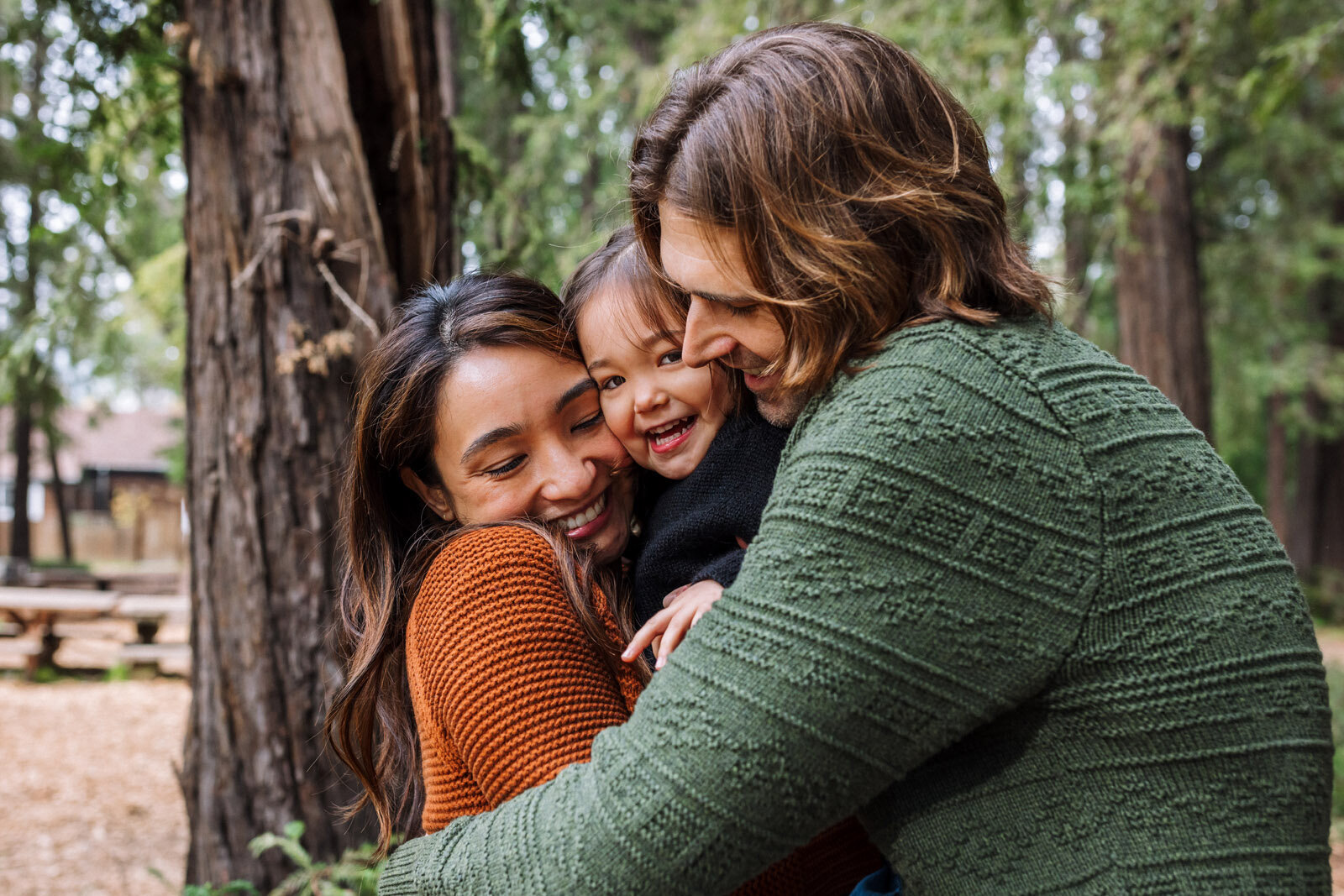 mom and dad snuggling toddler daughter they are holding outdoors as she looks at the camera and smiles