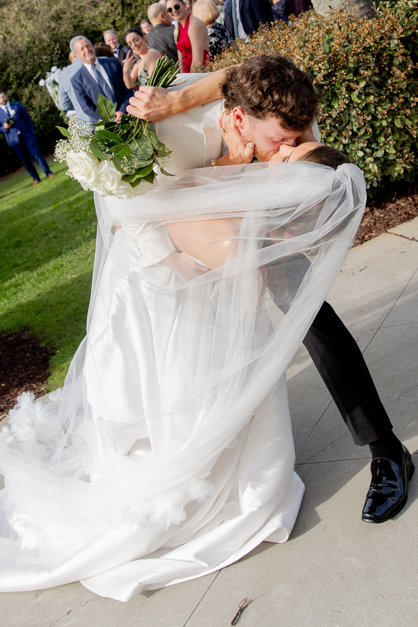 A cropped photo of a groom dipping and kissing his bride on their wedding day outside. The photo feels editorial and luxe.