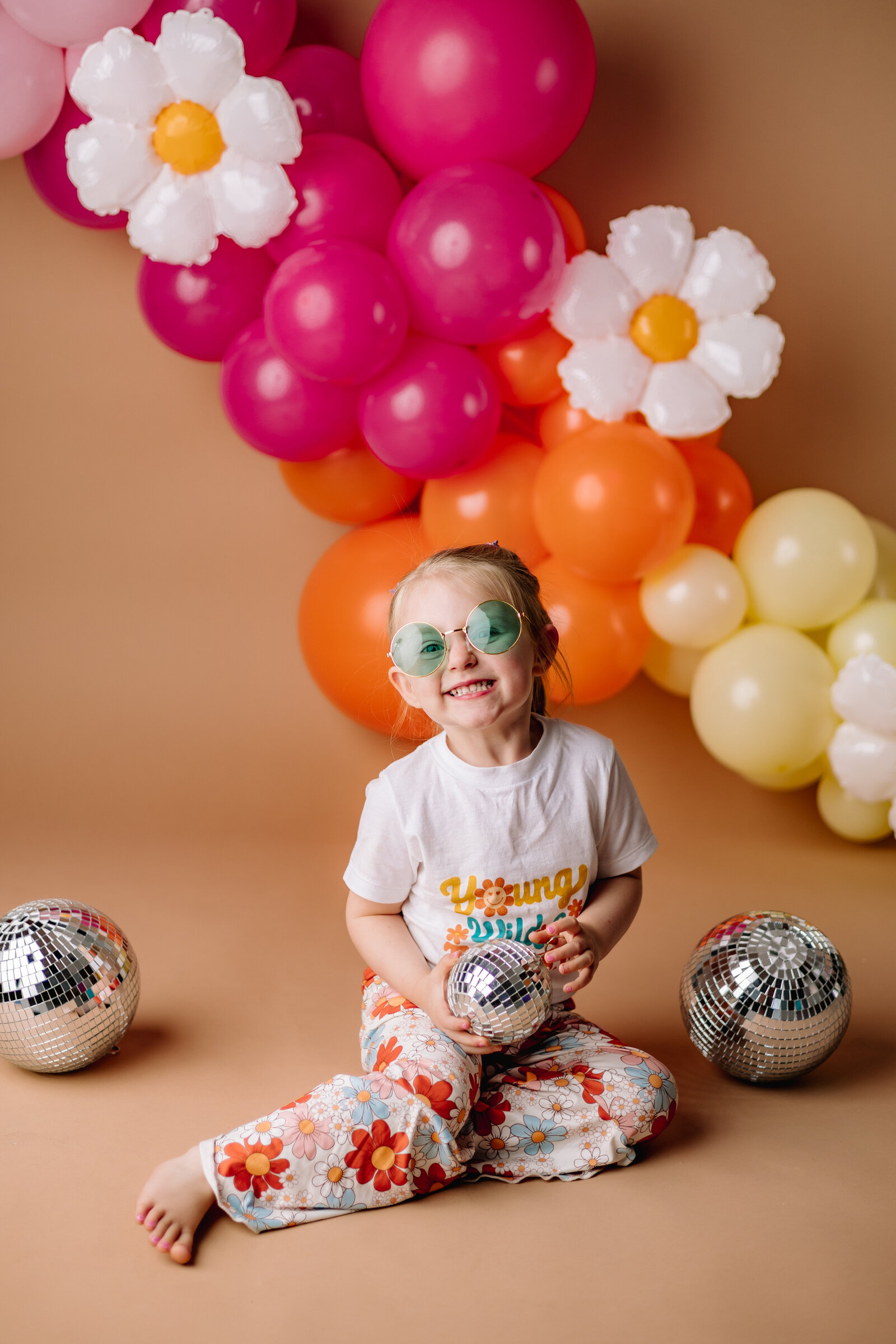 Sweet little girl with round sunglasses, smiles joyfully at the camera surrounded by balloons and disco balls