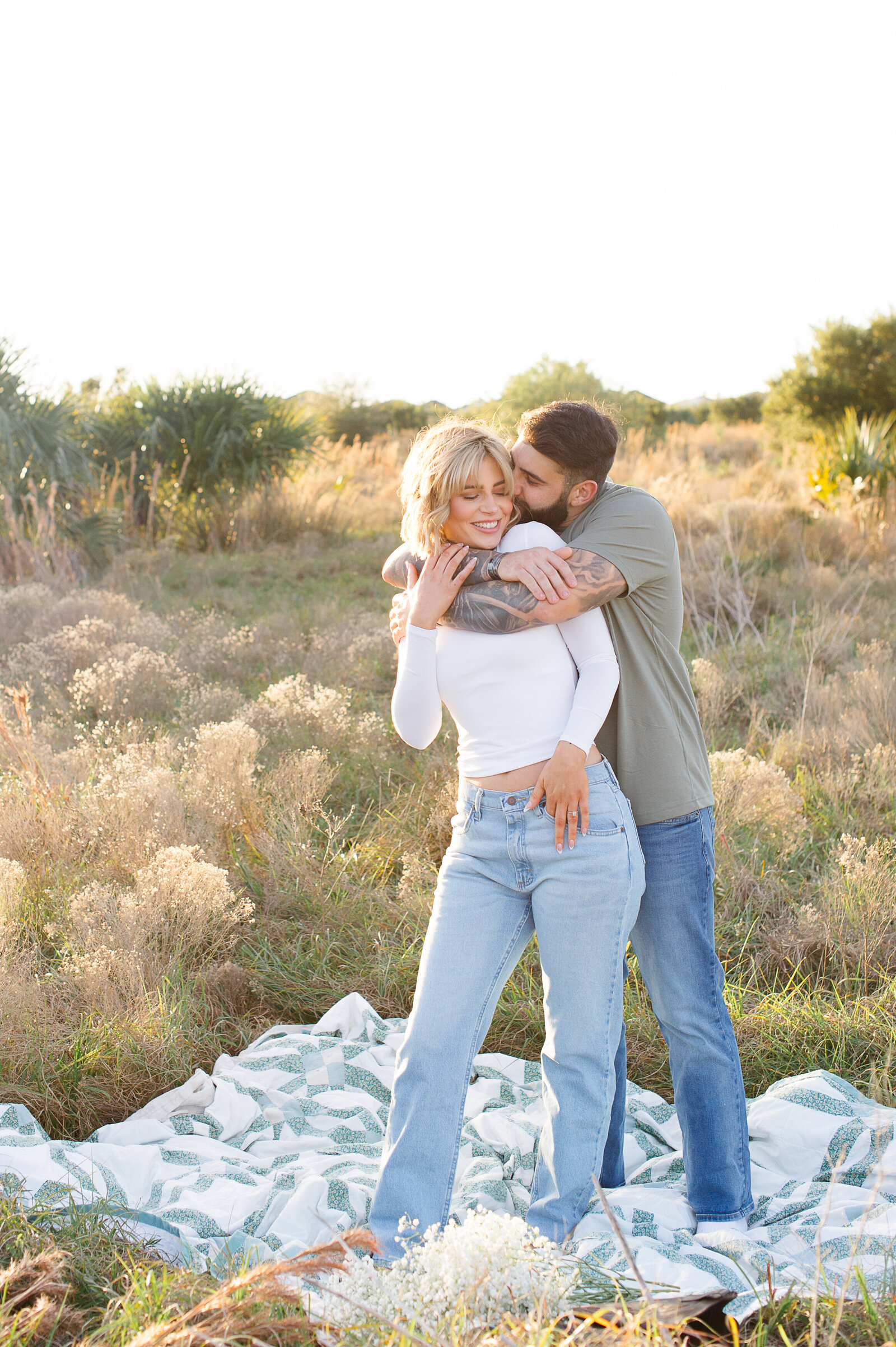 Newly engaged couple standing in a. field of tall pampas on a quilt while the man hugs the woman from behind and whispers in her ear