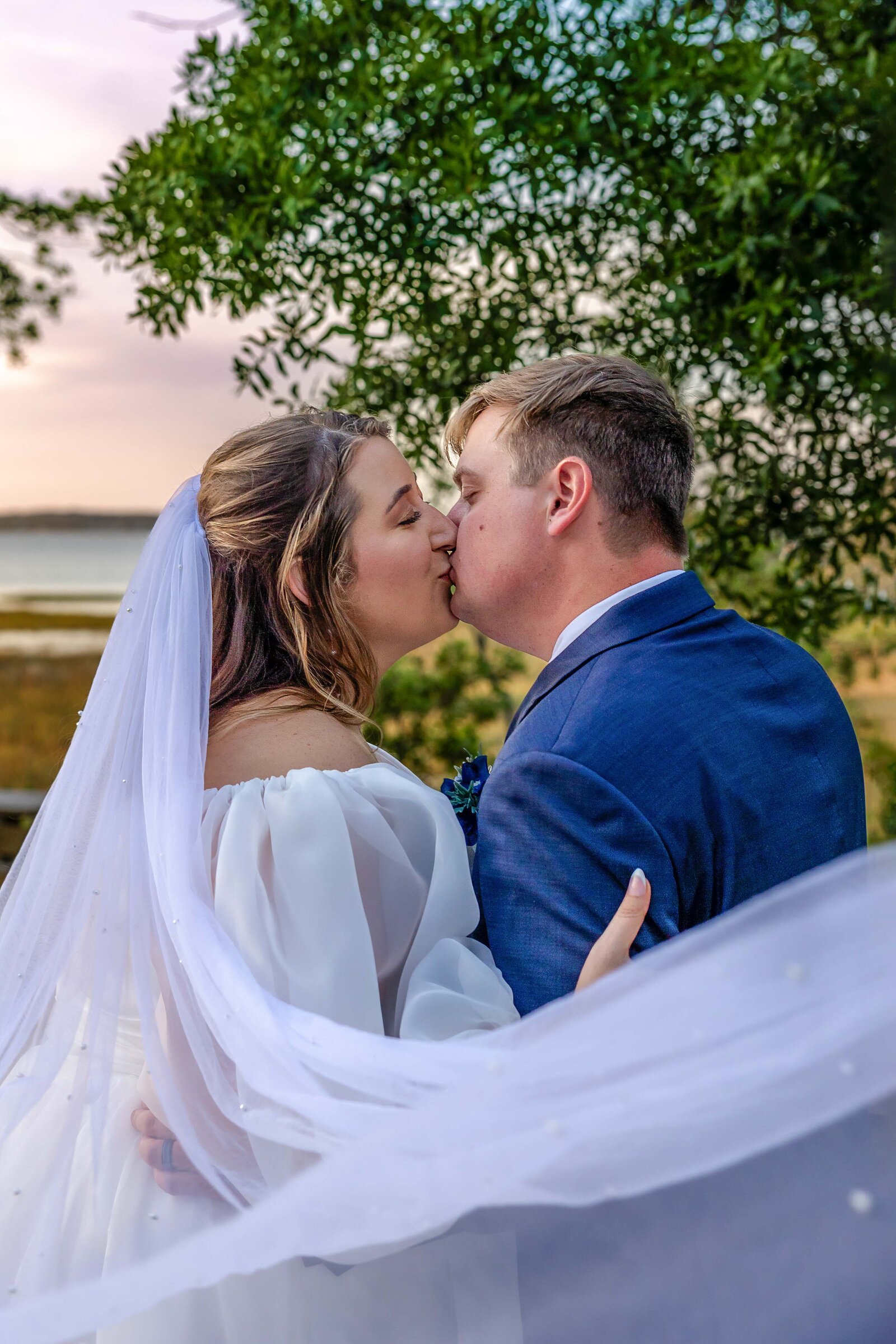 The bride and groom share a kiss near a marsh with the bride's veil flowing in the breeze, captured at sunset during a Pine Knoll Shores Aquarium, NC wedding, blending romance with natural beauty.