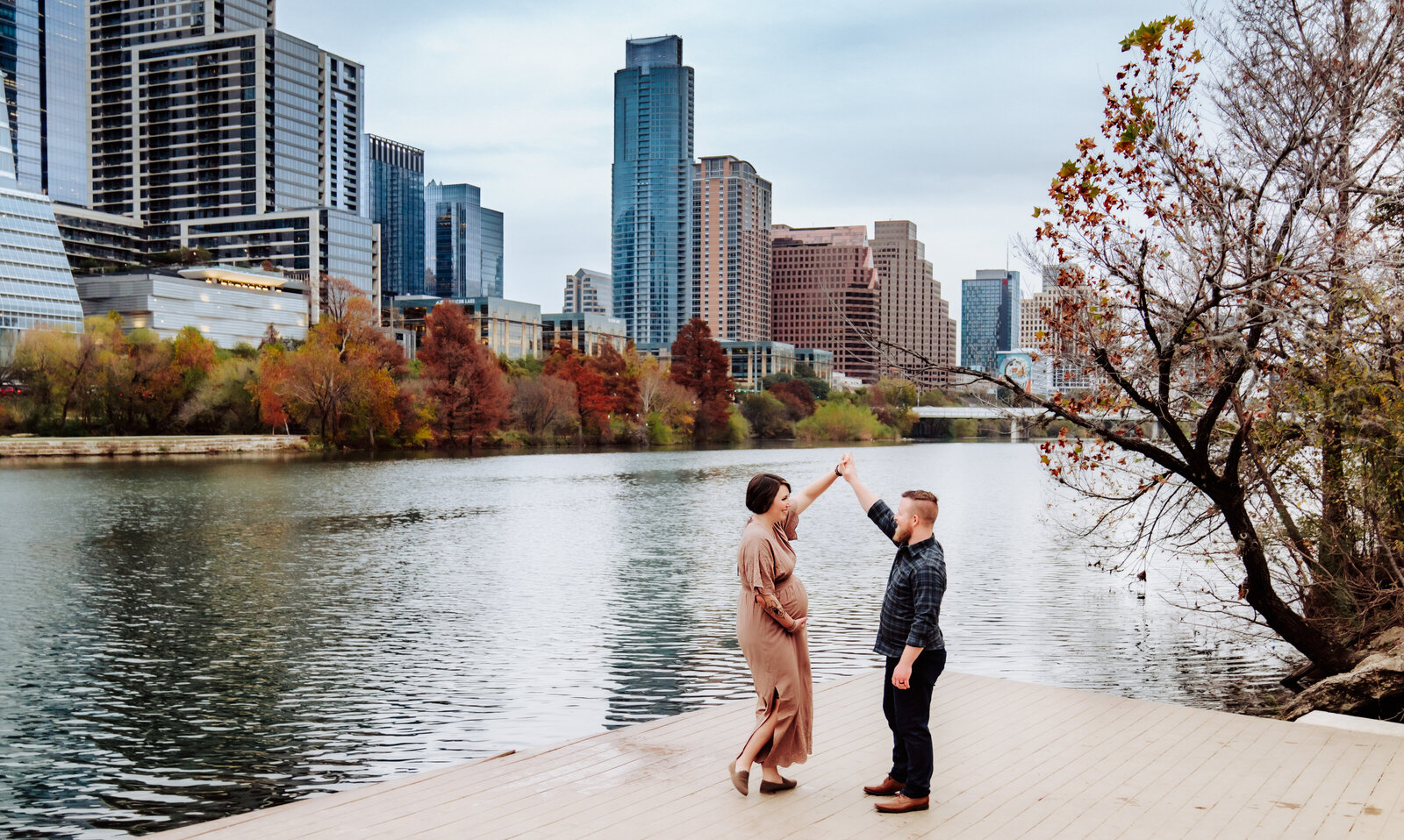 Maternity Photographer, a man and woman dance on a platform near the river, cityscape just across the water