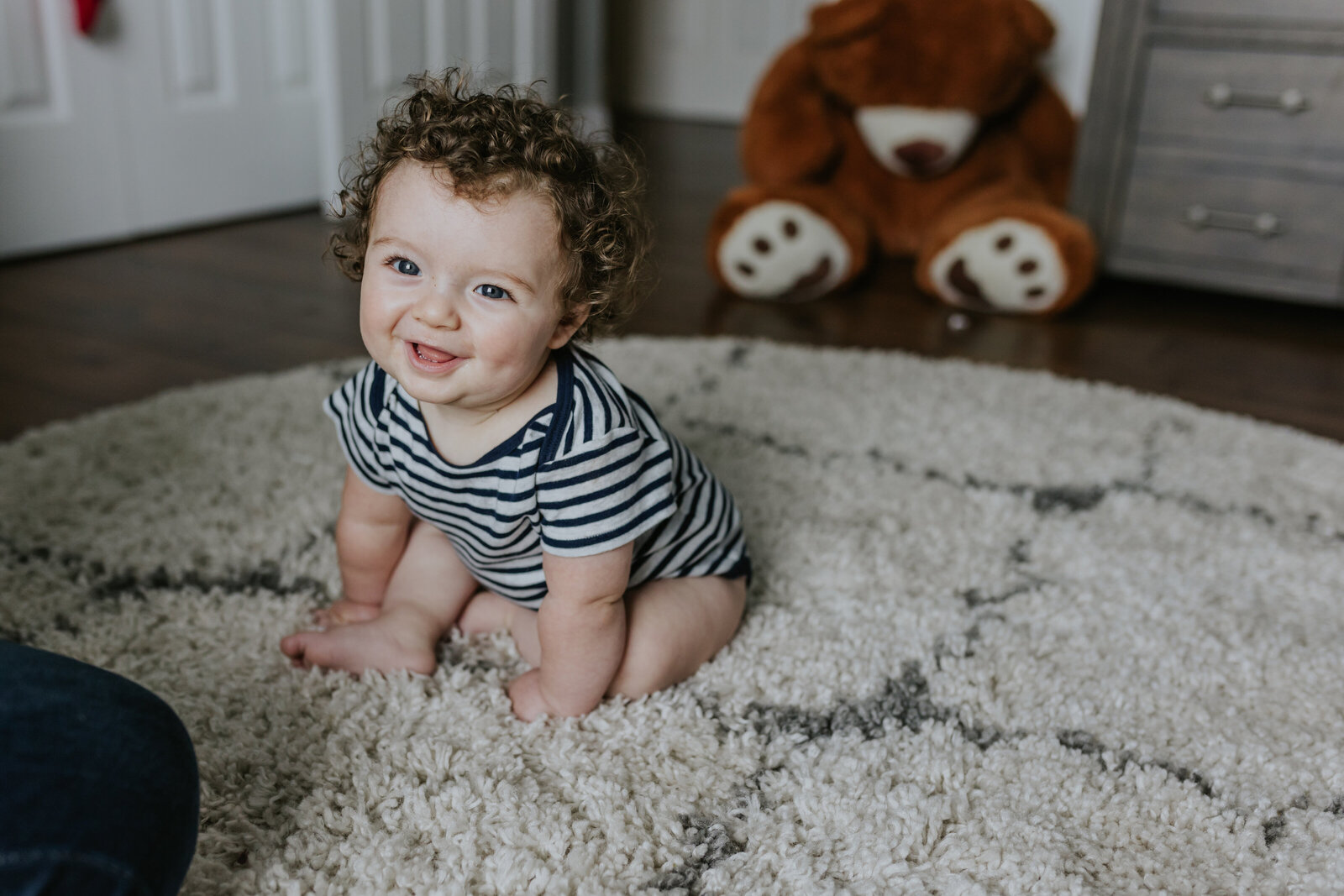 A baby sits on a shag rug and smiles at the camera