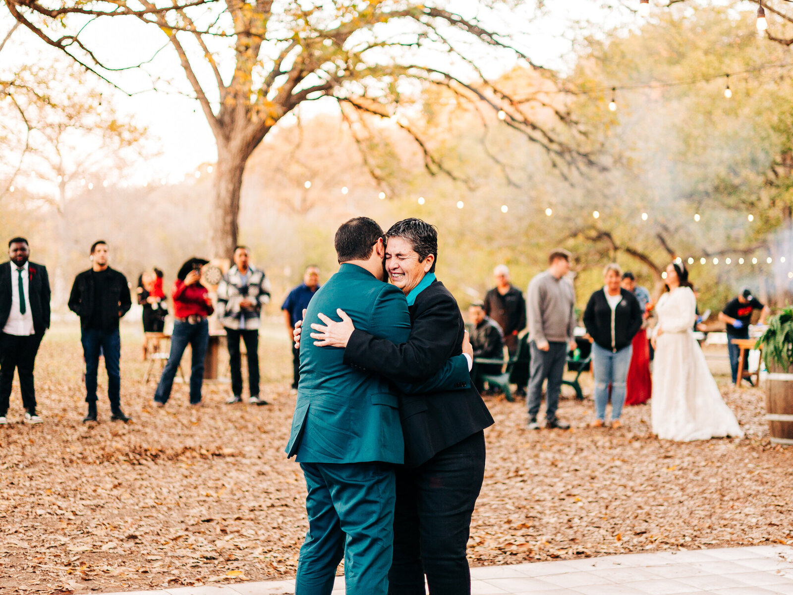 Two people embrace and dance together outdoors on a leaf-covered ground, surrounded by onlookers. String lights twinkle overhead, autumn trees fill the background, and a person in a white dress stands to the right—captured by an Eau Claire Wedding Photographer.