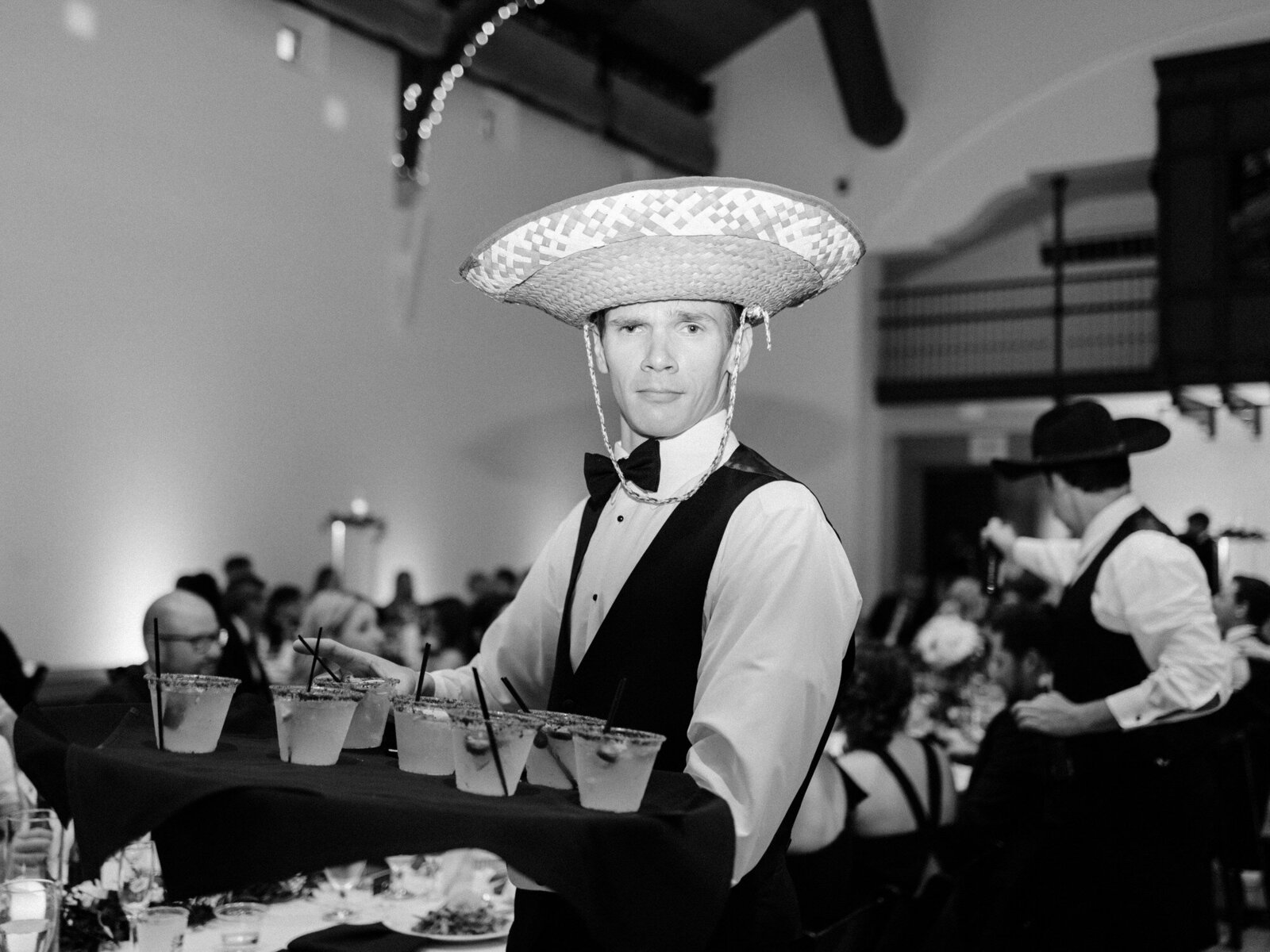 One of the groomsmen walks through the reception hall carrying a tray of drinks, wearing a sombrero. He is looking straight into the camera.