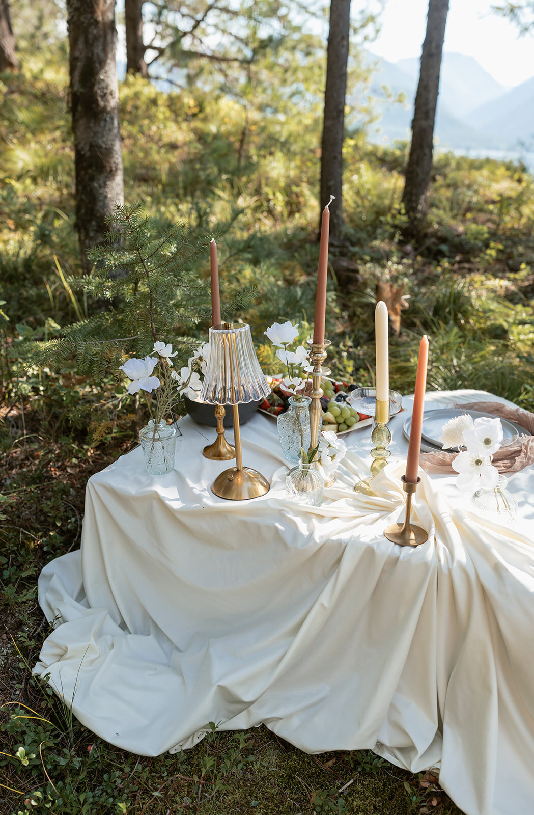 Elegant picnic-style elopement table styled with draped linens, gold candlesticks, and neutral florals in a forest setting, blending luxury with nature.