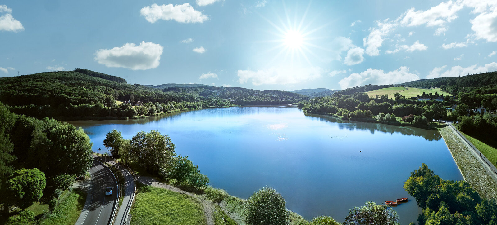 Panoramaaufnahme eines klaren Sees inmitten grüner Hügellandschaft bei Sonnenschein – Drohnenfotografie mit Weitwinkelperspektive.