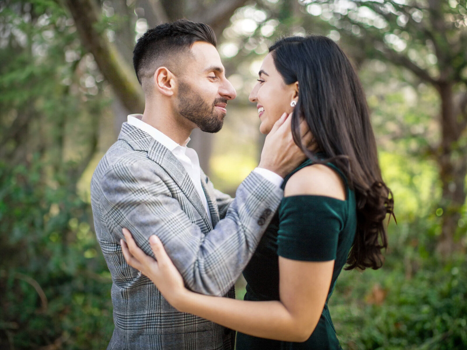 A couple stands closely in a lush, green forest. The man, in a plaid suit, gently holds the womans face as they smile at each other. The woman wears a dark green, off-the-shoulder dress, and they are surrounded by blurred tree branches.