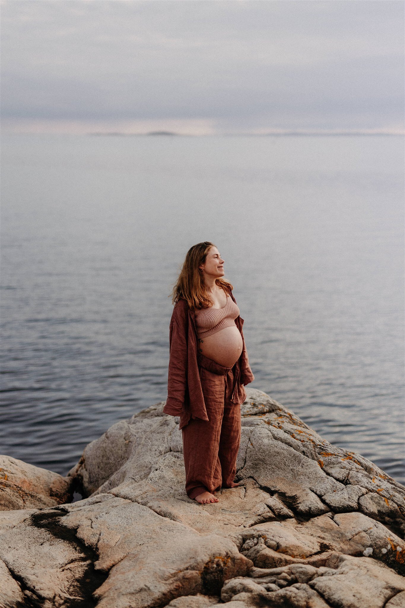 på höga klippor står en gravid mamma iklädd rostbrunt linneset