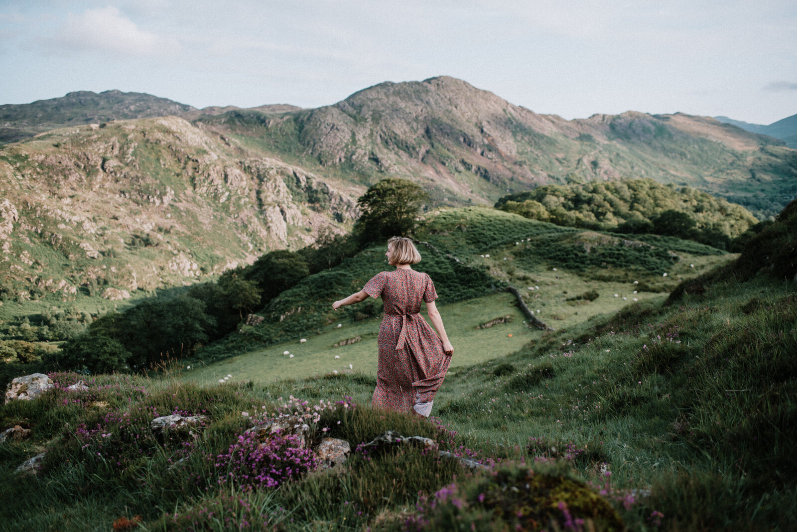 woman in pink floral dress is spinning atop a green mountain covered in wild flowers